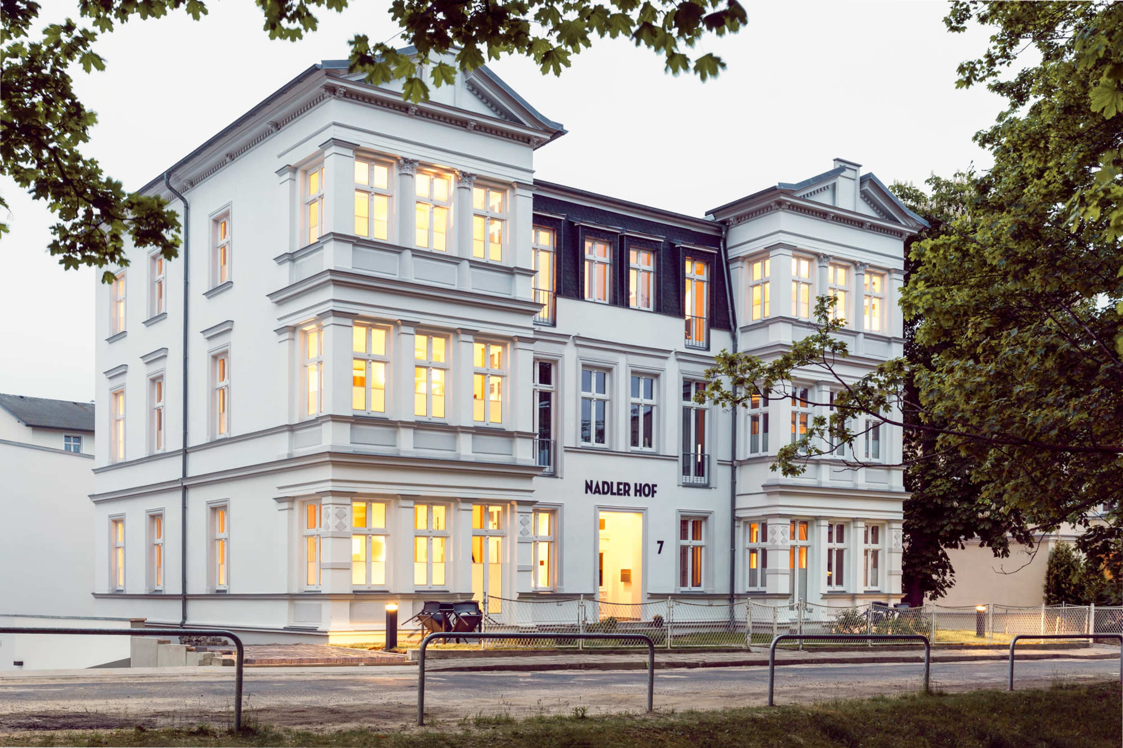 A large, white, multi-story building with numerous windows, illuminated from within, located at "Nadler Hof 7."
