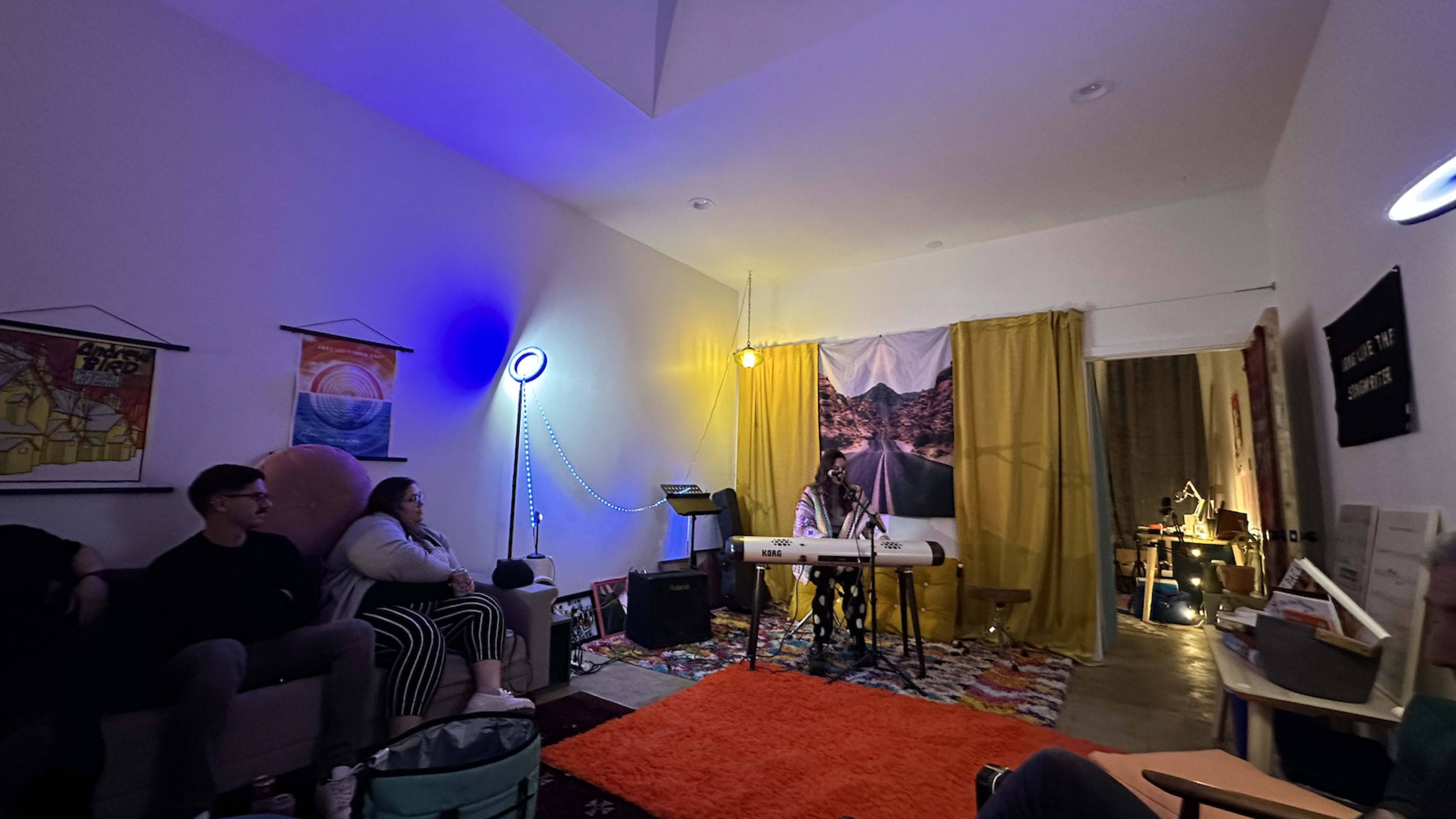 A musician performs at a small indoor gathering, with colorful lighting and a cozy seating arrangement.