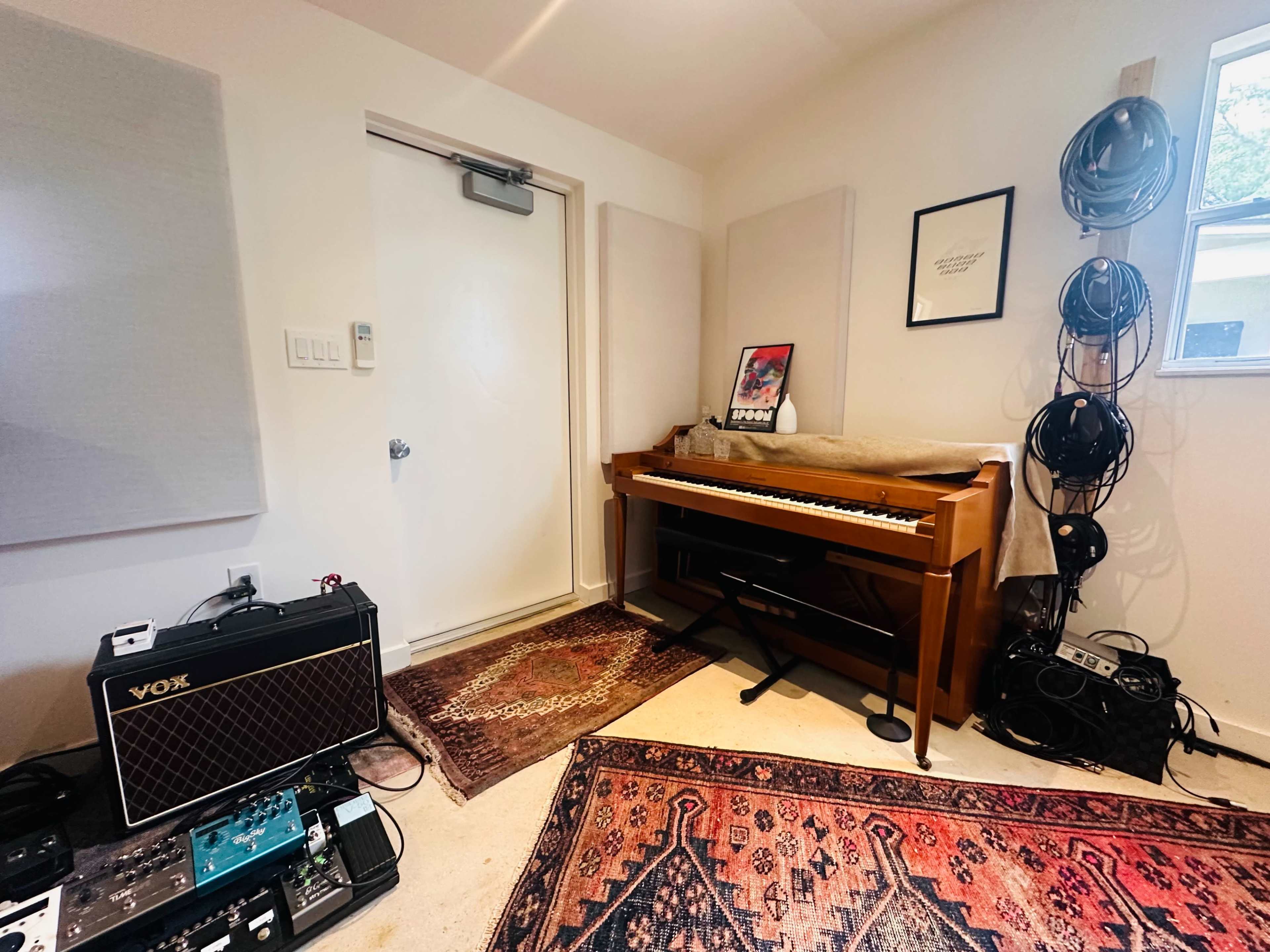 The image shows a small room with a piano against one wall, a door, and various musical equipment including pedals and cables on the floor.