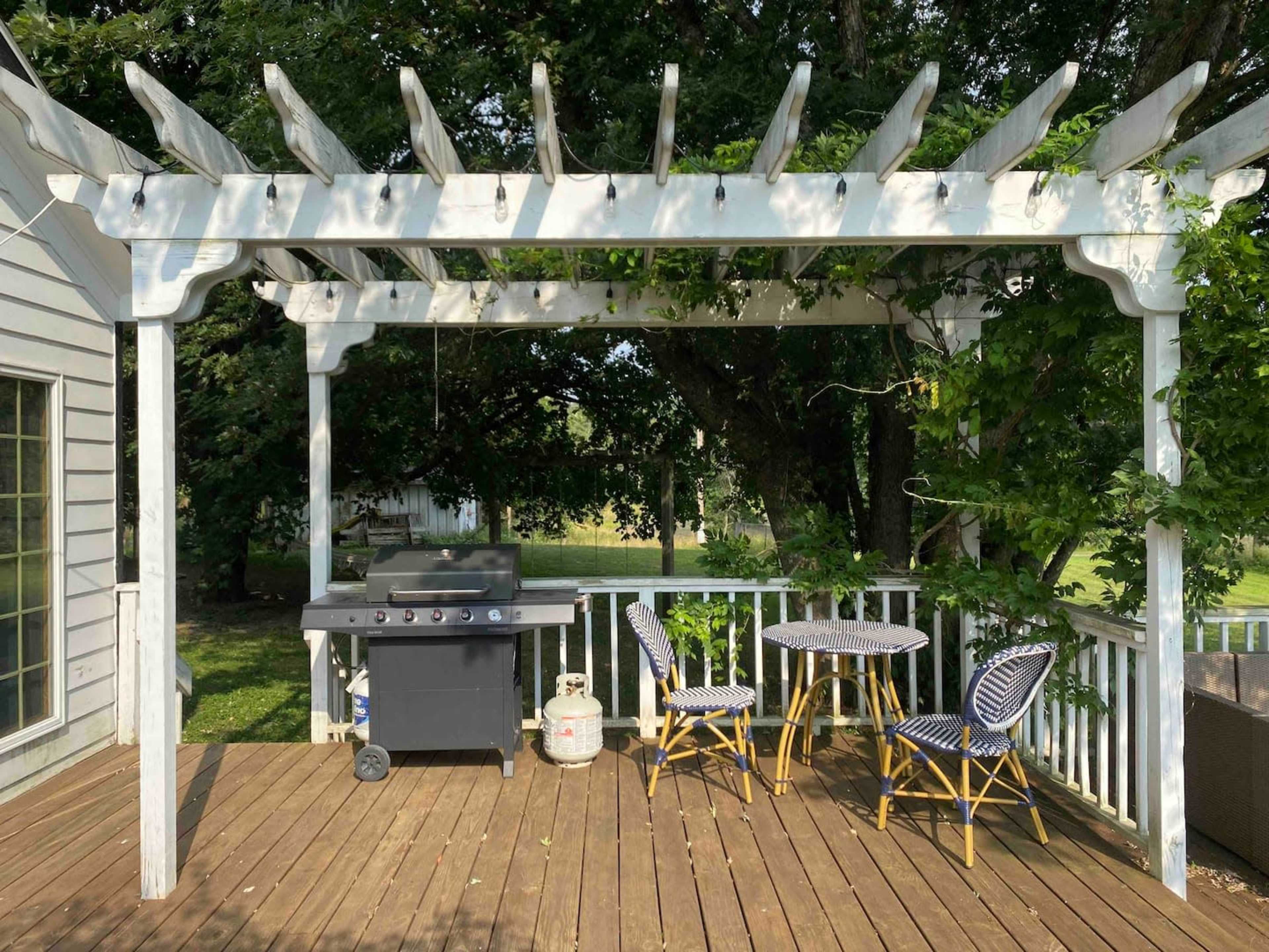 A wooden deck features a white pergola adorned with lights, a gas grill, and two bistro tables with chairs.