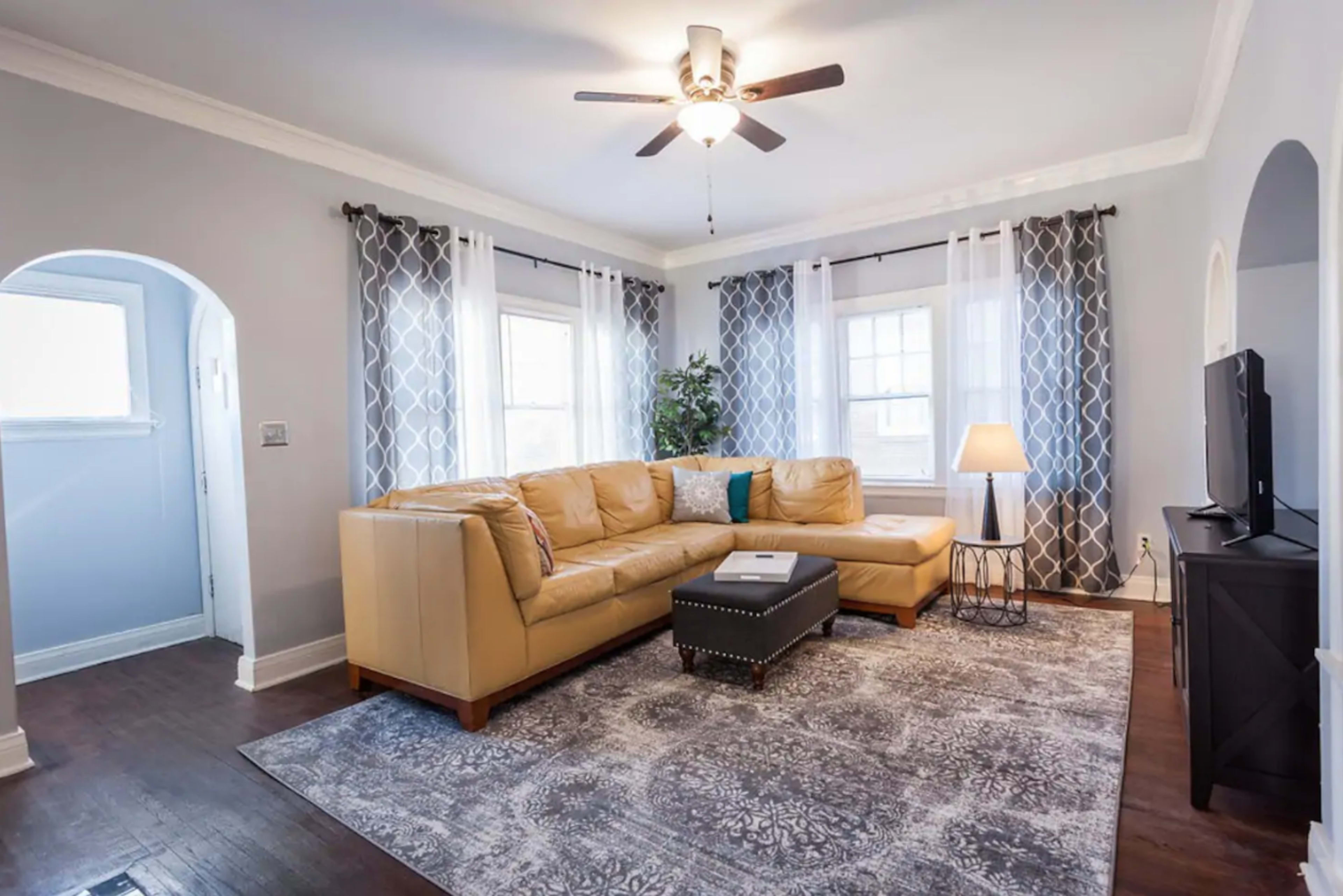 The image shows a living room with a tan sectional couch, a coffee table, large windows with patterned curtains, and a ceiling fan.