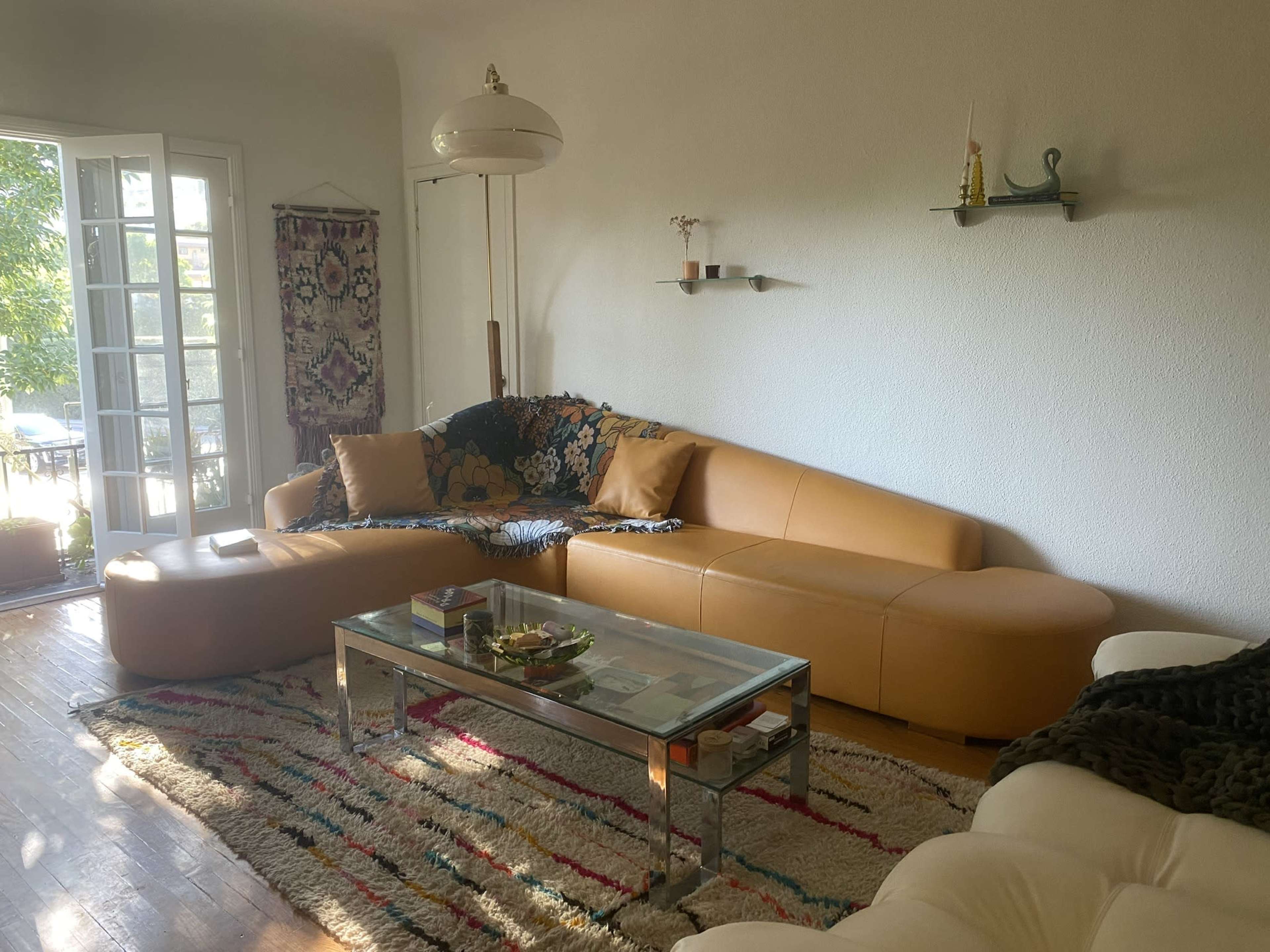 The living room features a curved leather sofa, a glass coffee table with decorative items, and large windows that open to a view of greenery outside.