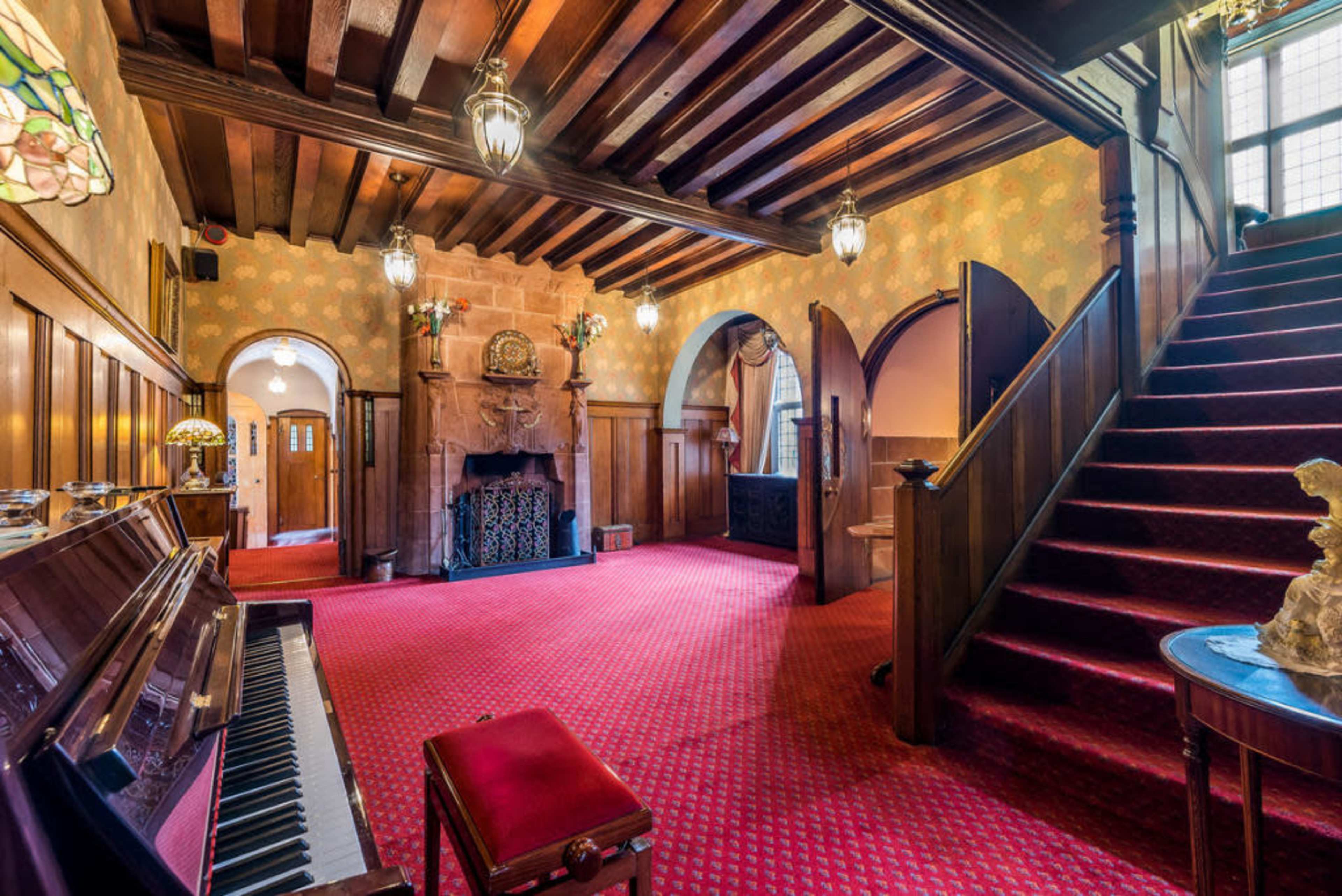 An elegant foyer with wooden beams, a grand staircase, a piano, and arched doorways leading to other rooms.