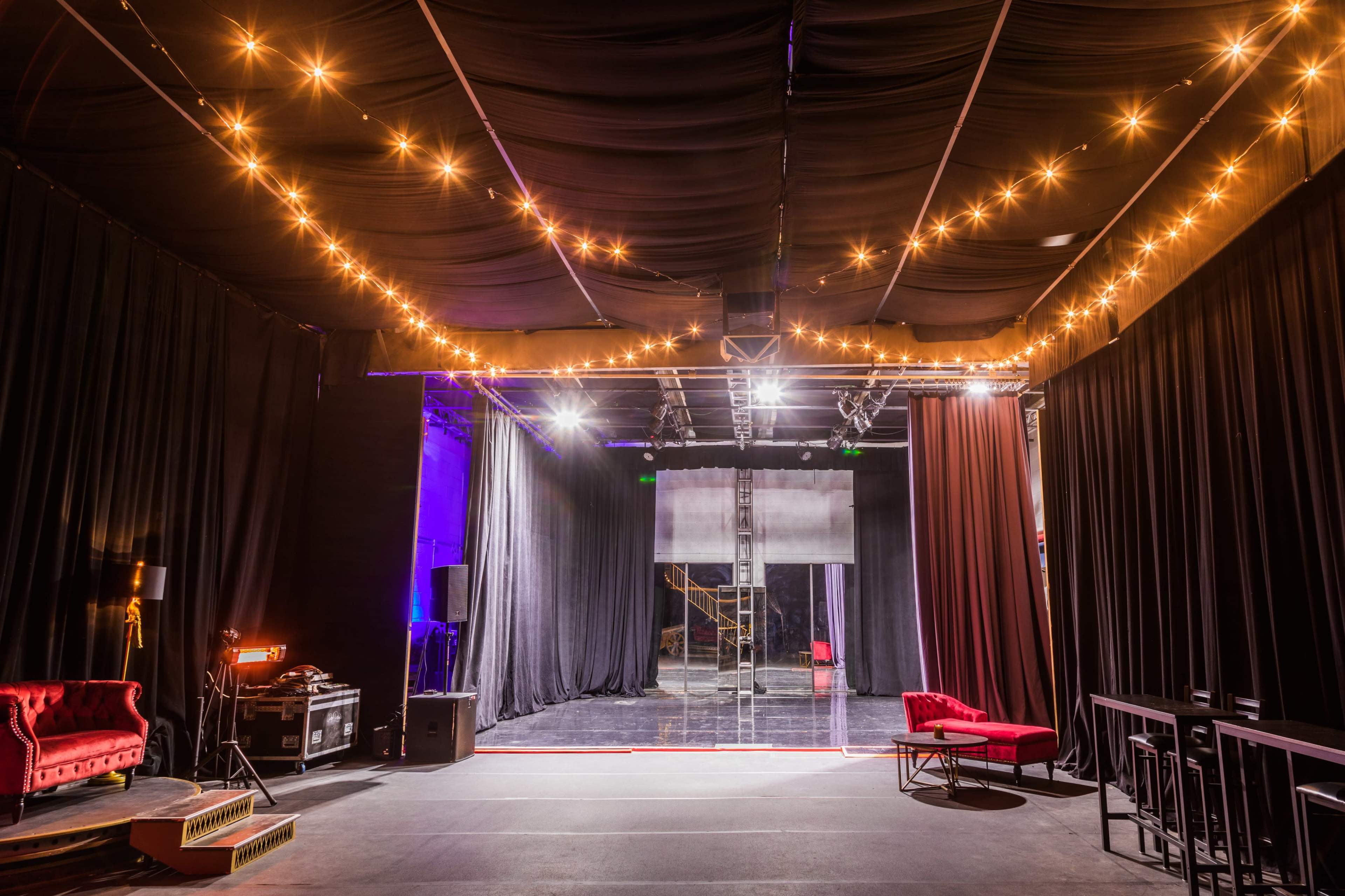 The image shows an empty stage with soft lighting and two red sofas set against black drapery.