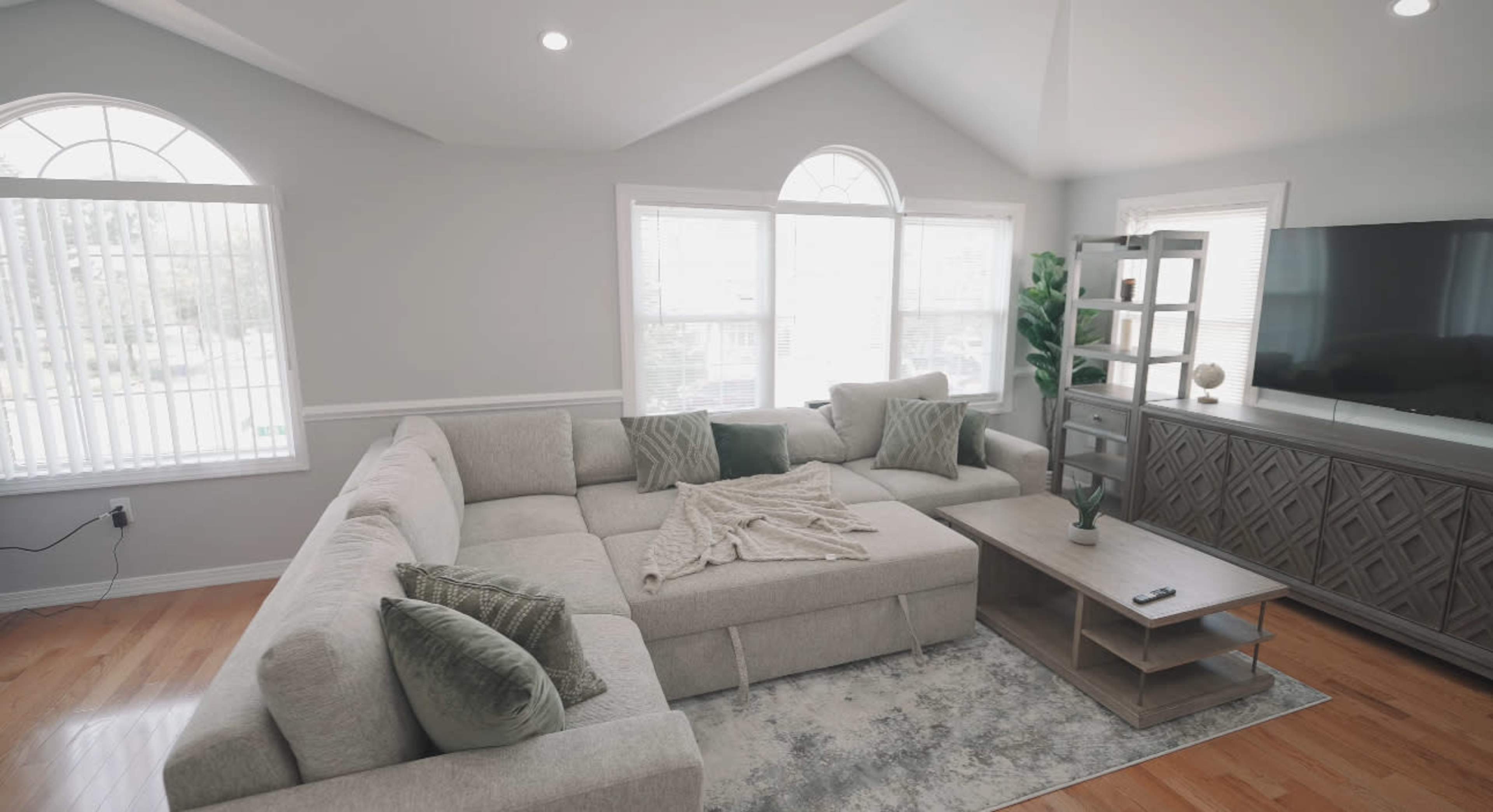 The image shows a spacious living room featuring a large sectional sofa, a light-colored area rug, and a television stand against the wall.