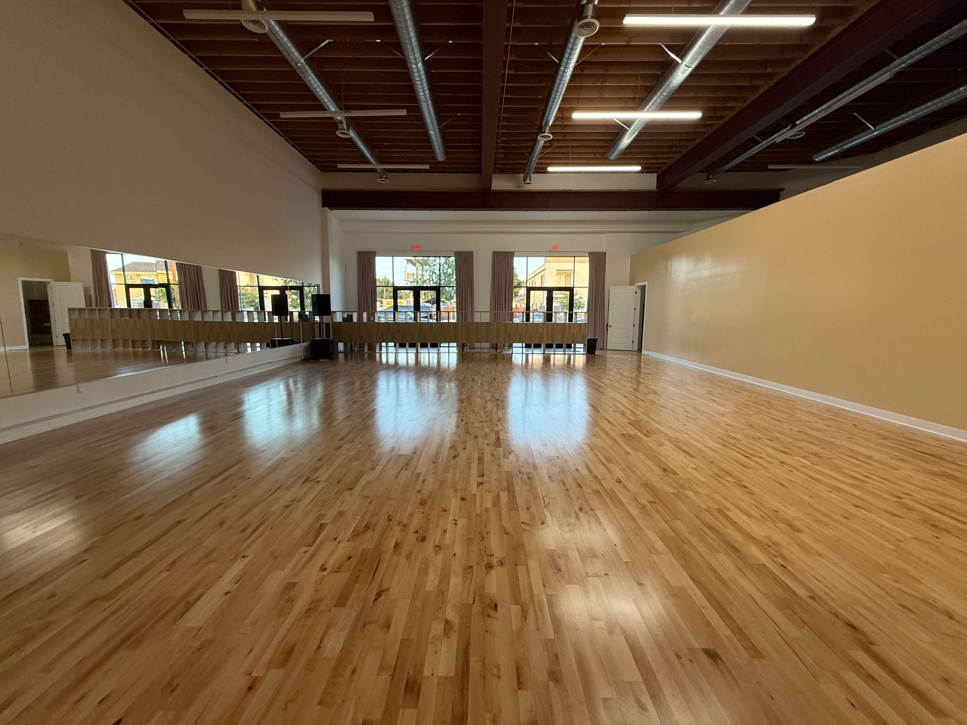 The image shows a spacious wooden dance studio with large mirrors on one wall and bright natural light coming through multiple windows.