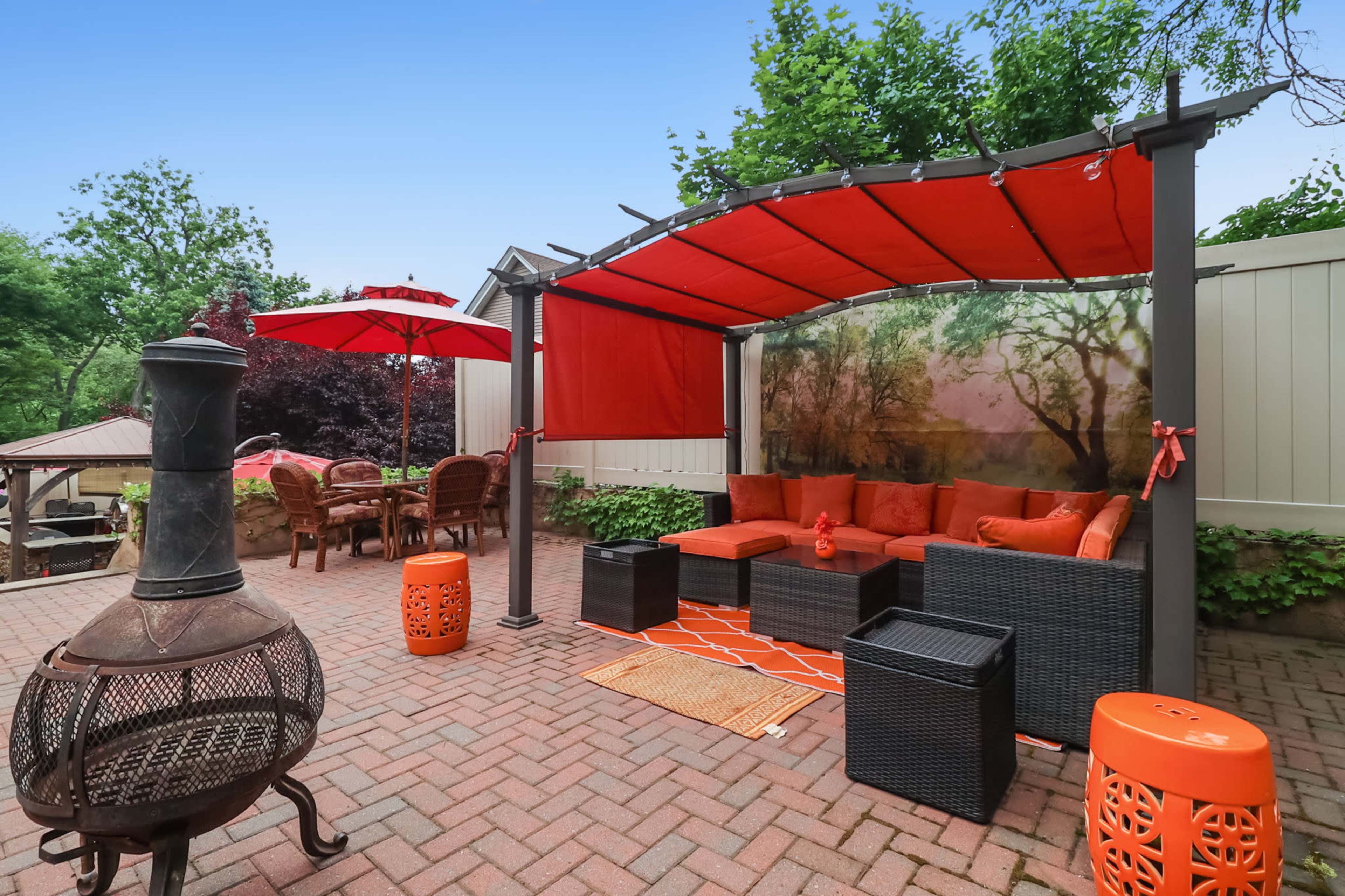 The patio features a covered seating area with orange cushions, a matching rug, a fire pit, and a dining set beneath an umbrella.