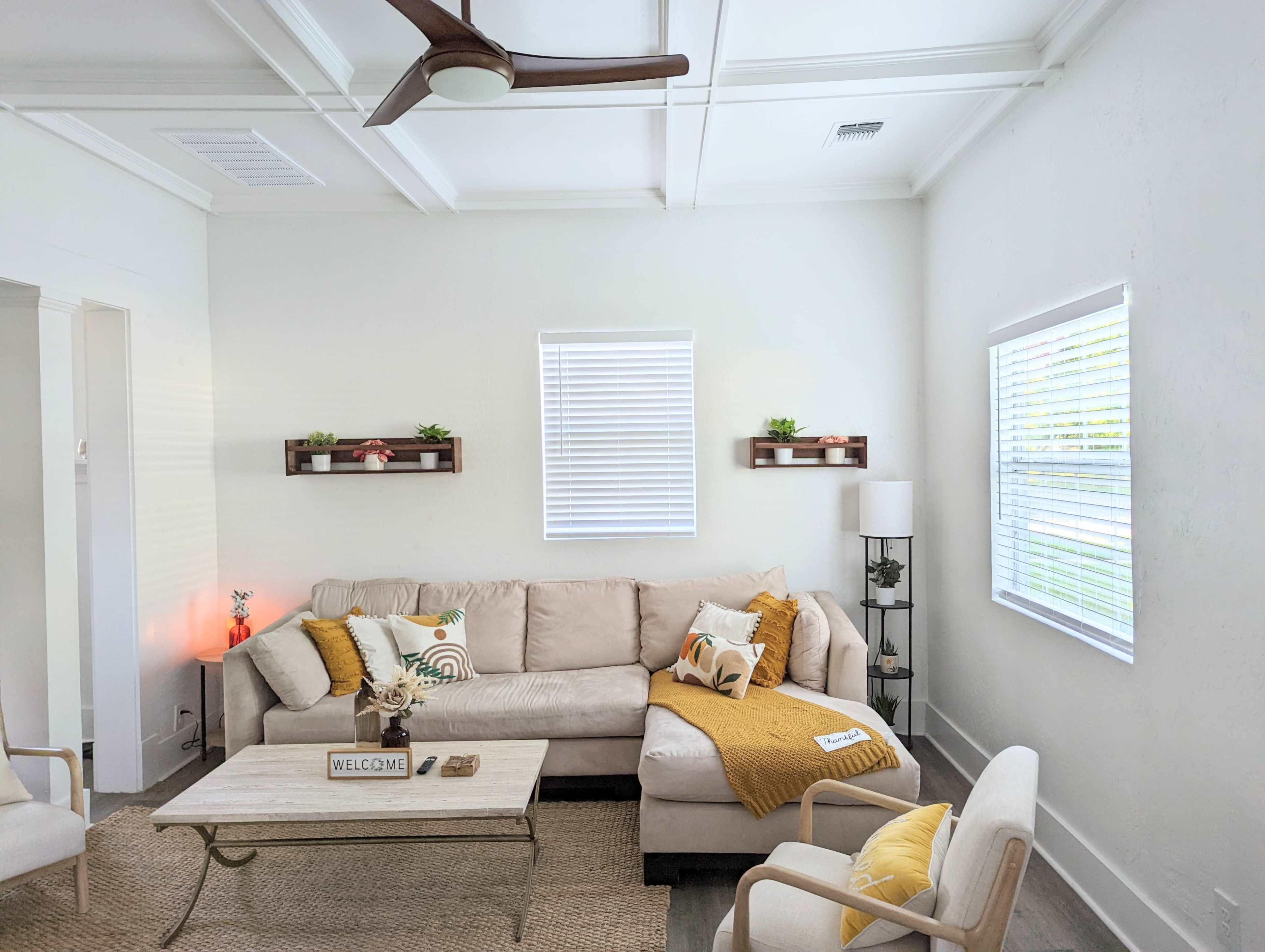 A light-filled living room features a beige sectional sofa, a coffee table, a floor lamp, and shelves with small plants.