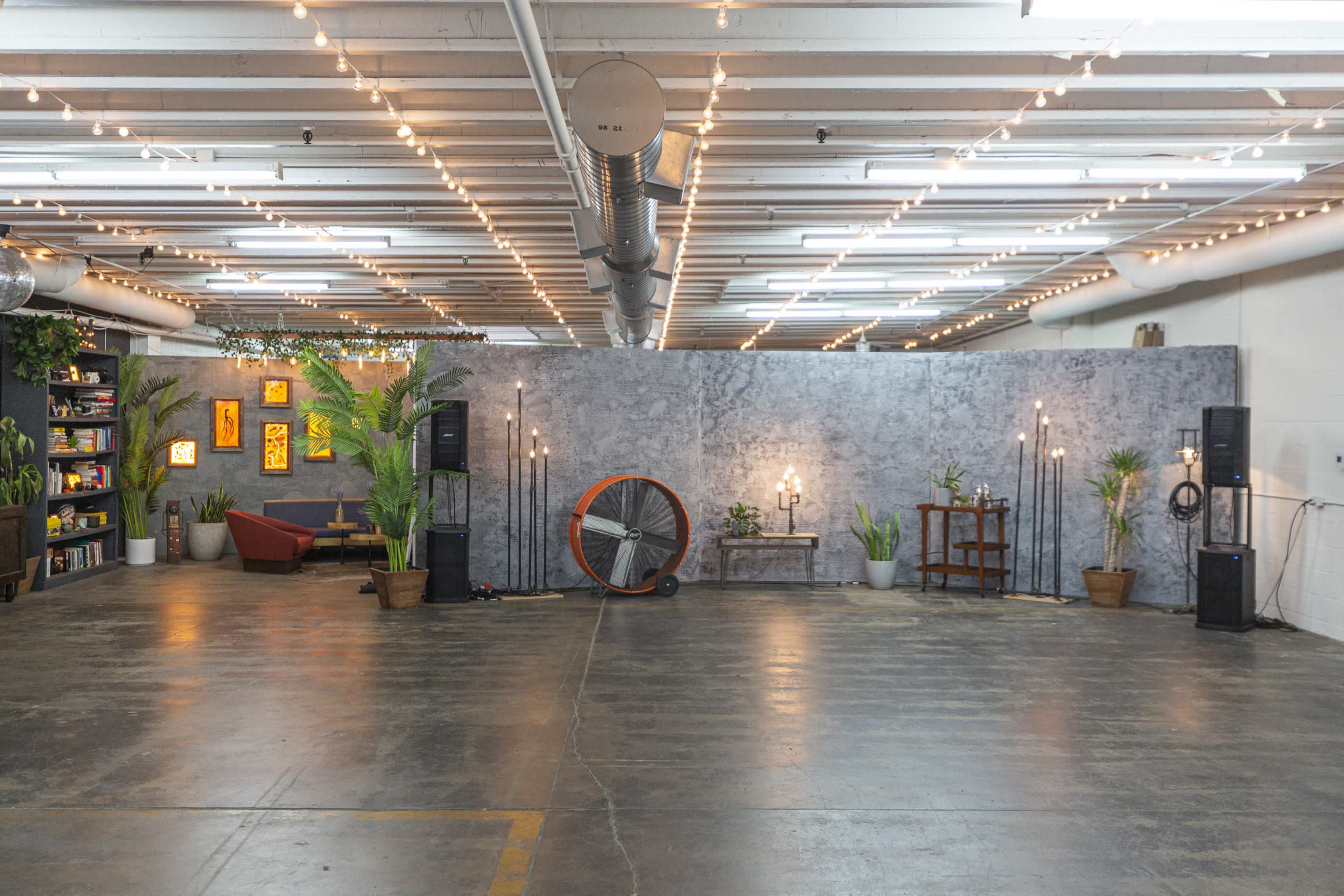 A spacious indoor event area with a gray backdrop, various potted plants, decorative lights, and audio equipment arranged throughout.