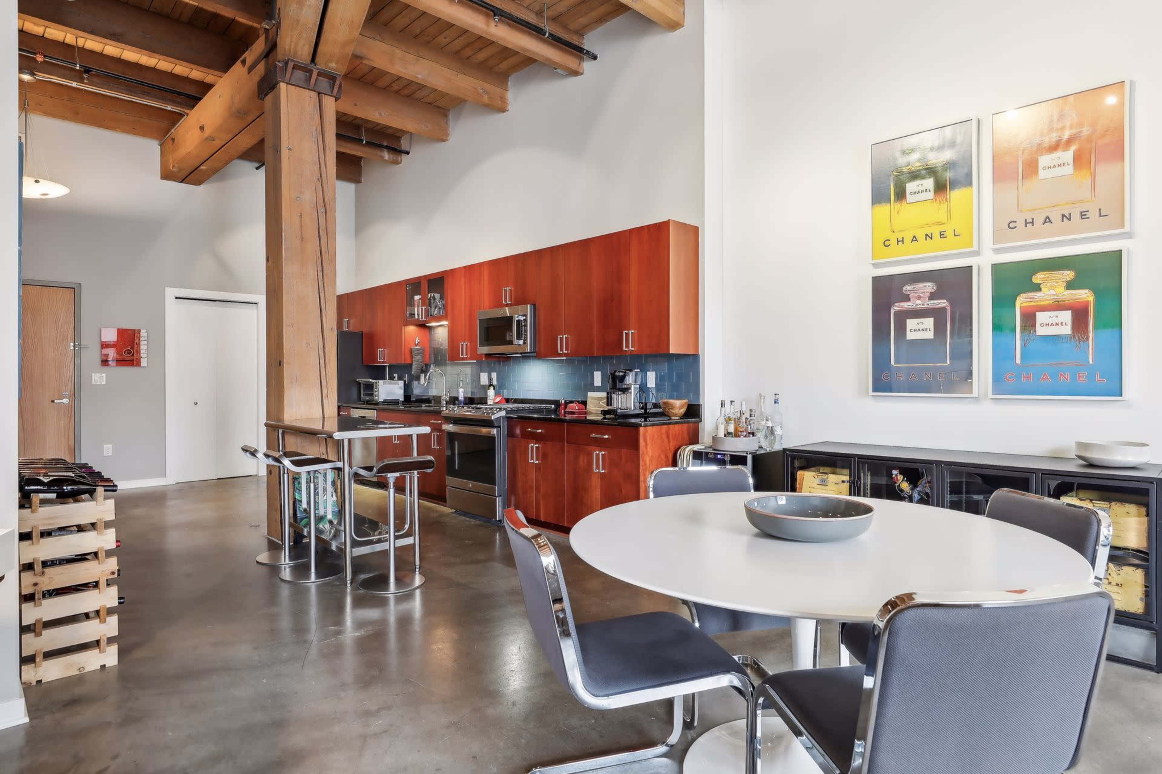 The image shows a modern kitchen and dining area featuring wooden beams, stainless steel appliances, a round dining table, and framed Chanel artworks on the wall.