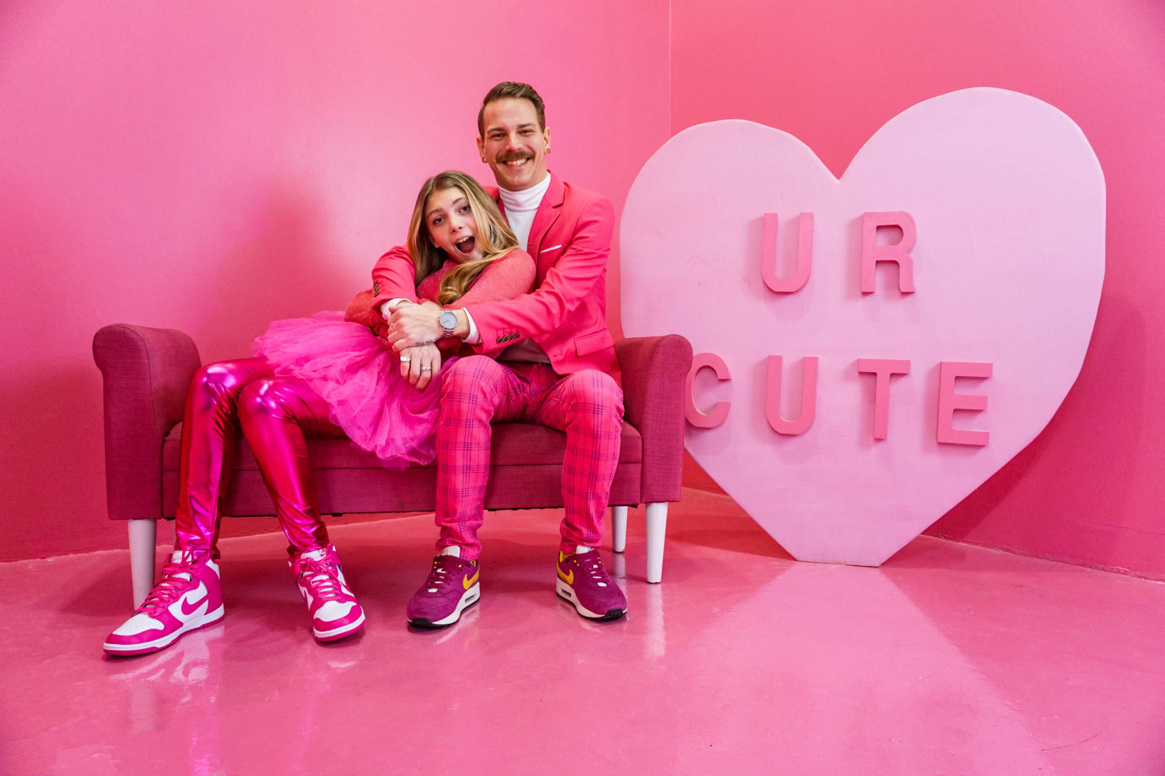 A man and a girl sit on a pink couch in a room decorated with pink walls and a large heart that says "UR CUTE."