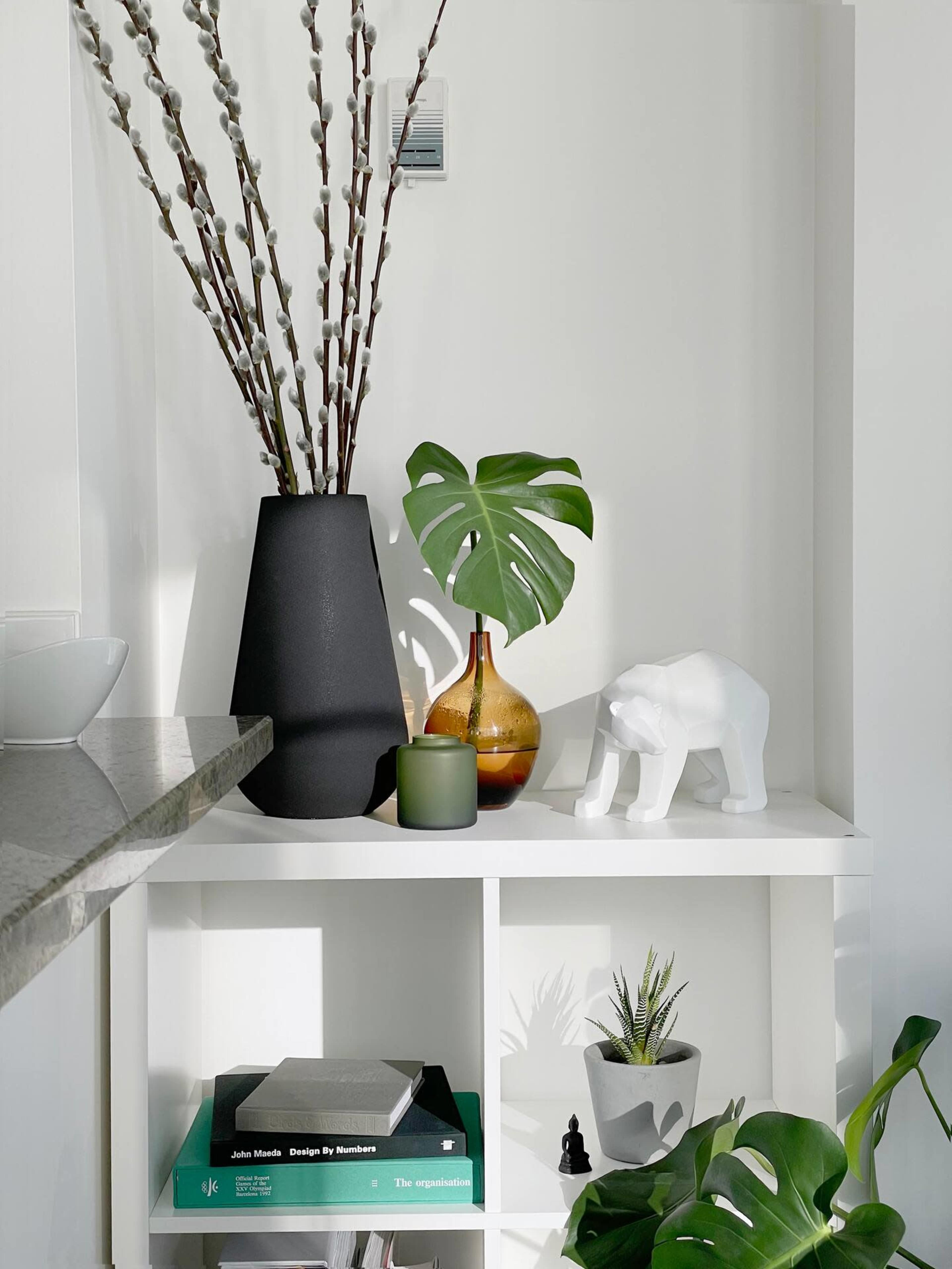 A minimalist shelf displays a black vase with branches, a brown glass bottle, a green candle, a white bear sculpture, and various plants.