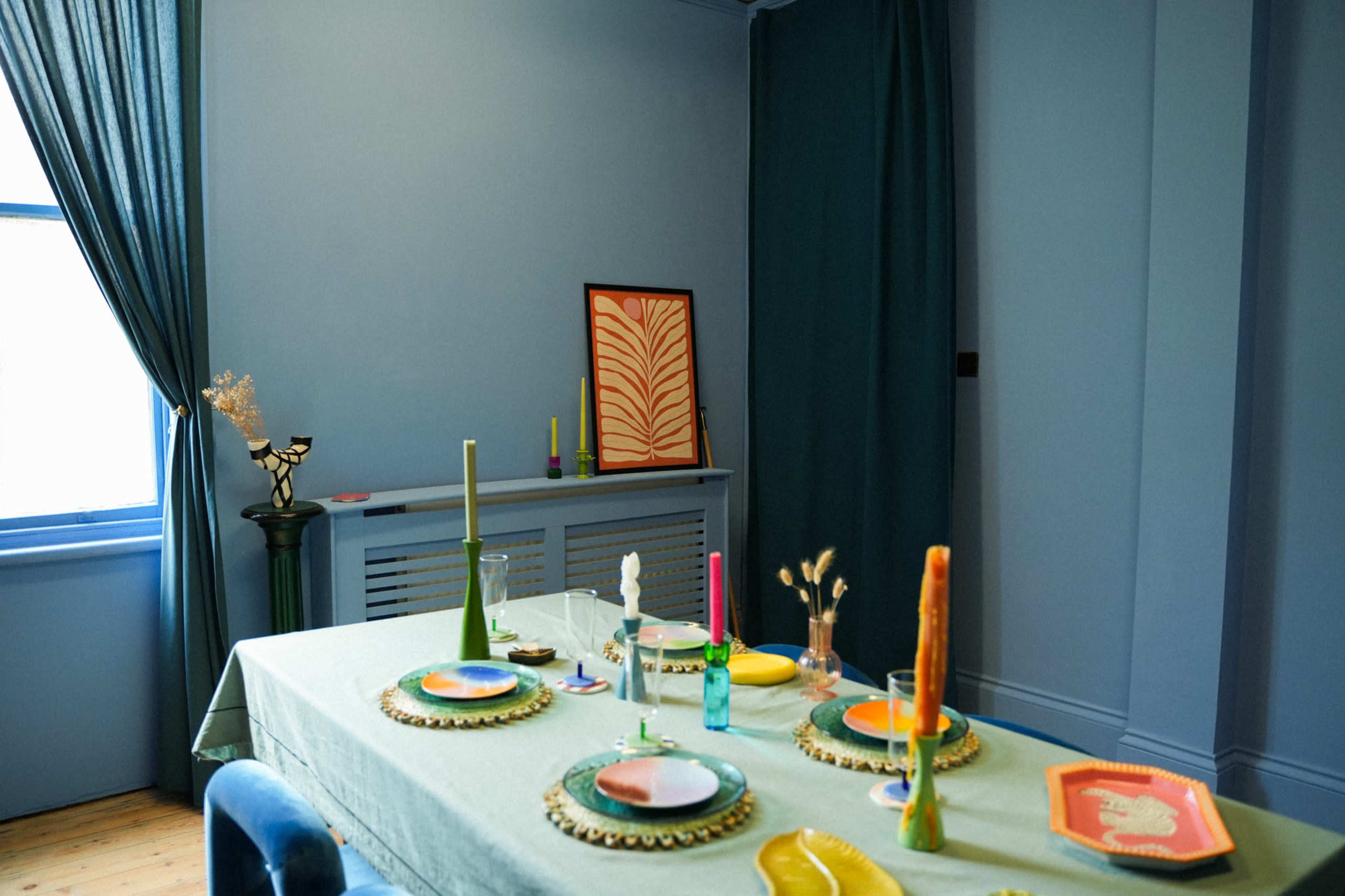 A dining table is set with colorful plates and candles in a blue-walled room featuring a window and decorative items.