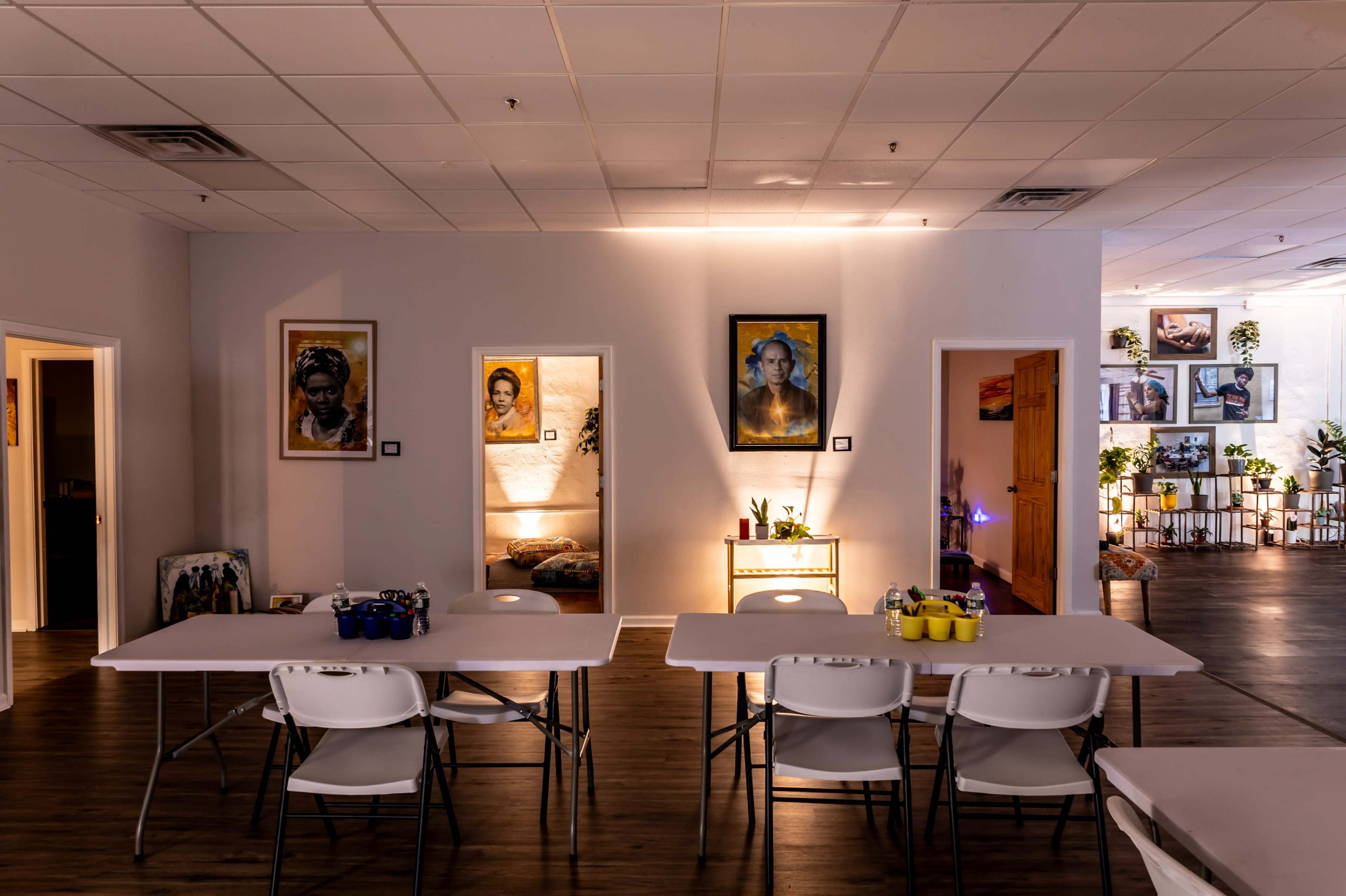 A spacious room features white tables and chairs, with artwork on the walls and plants visible in the background.