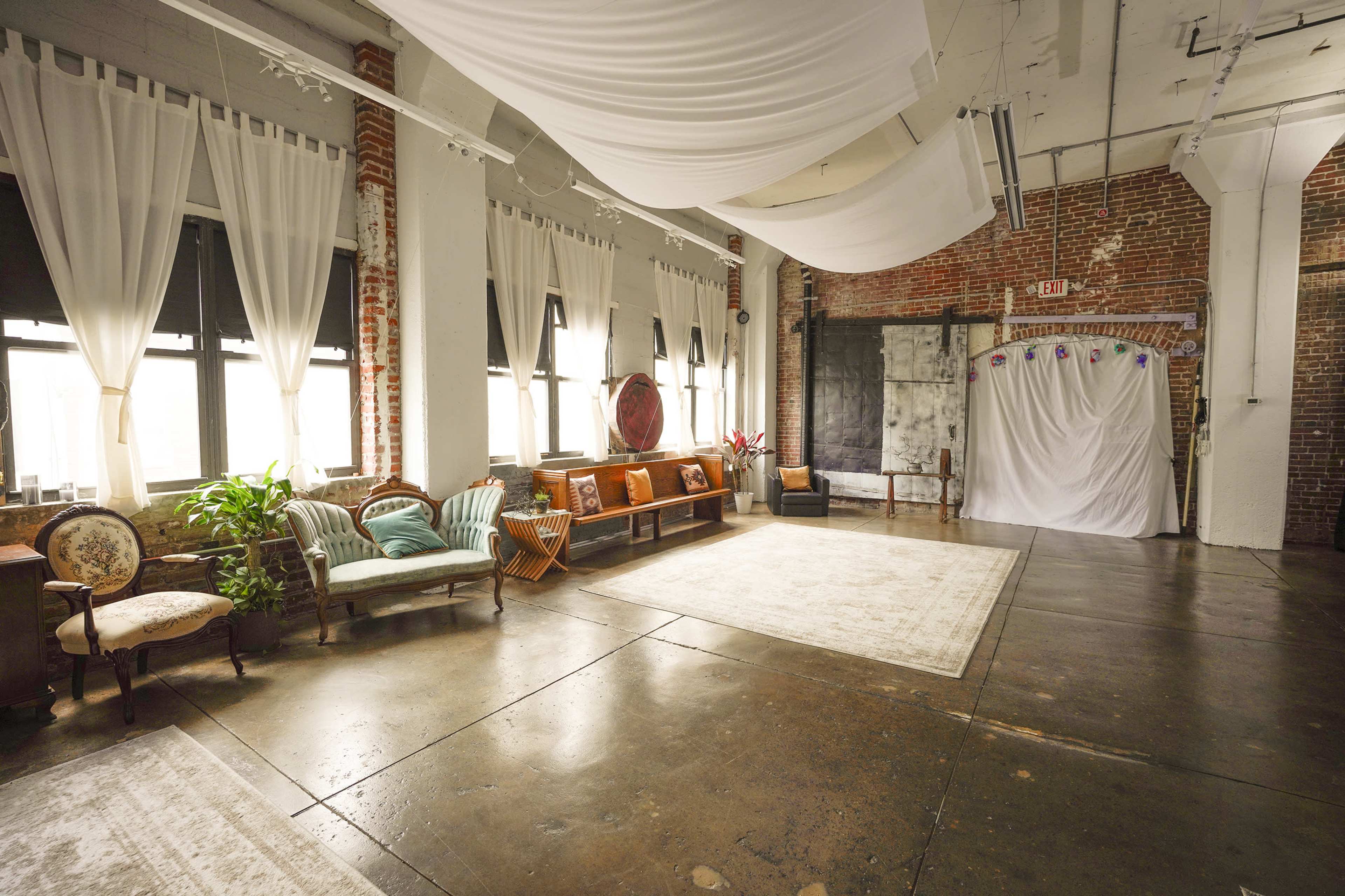 A spacious industrial room with large windows, a polished concrete floor, and a seating area featuring vintage furniture along one wall.