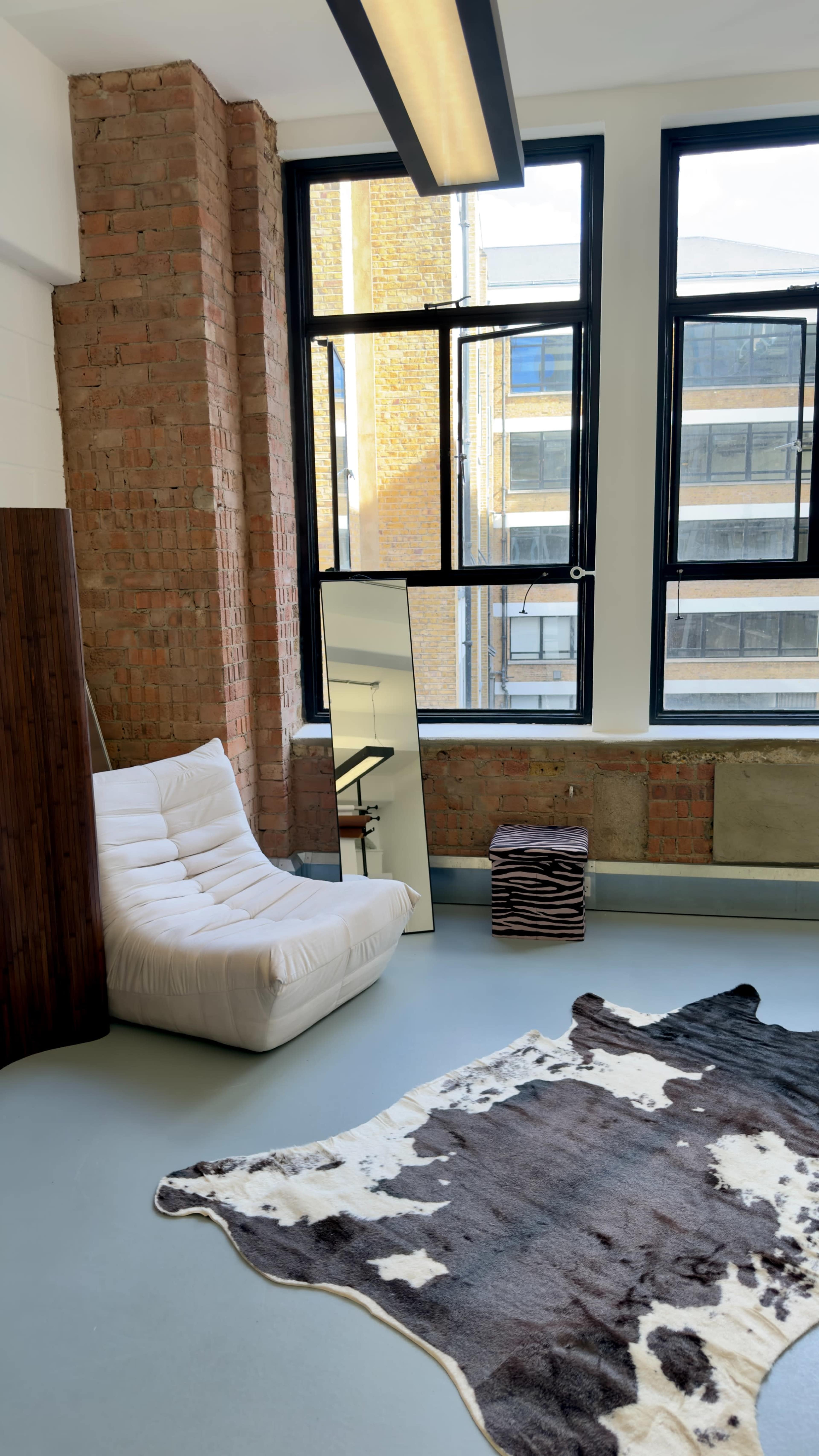 The image shows a modern indoor space featuring a white lounging chair, a large mirror, a stack of decorative boxes, and a patterned rug, with exposed brick walls and large windows allowing natural light to flood the room.