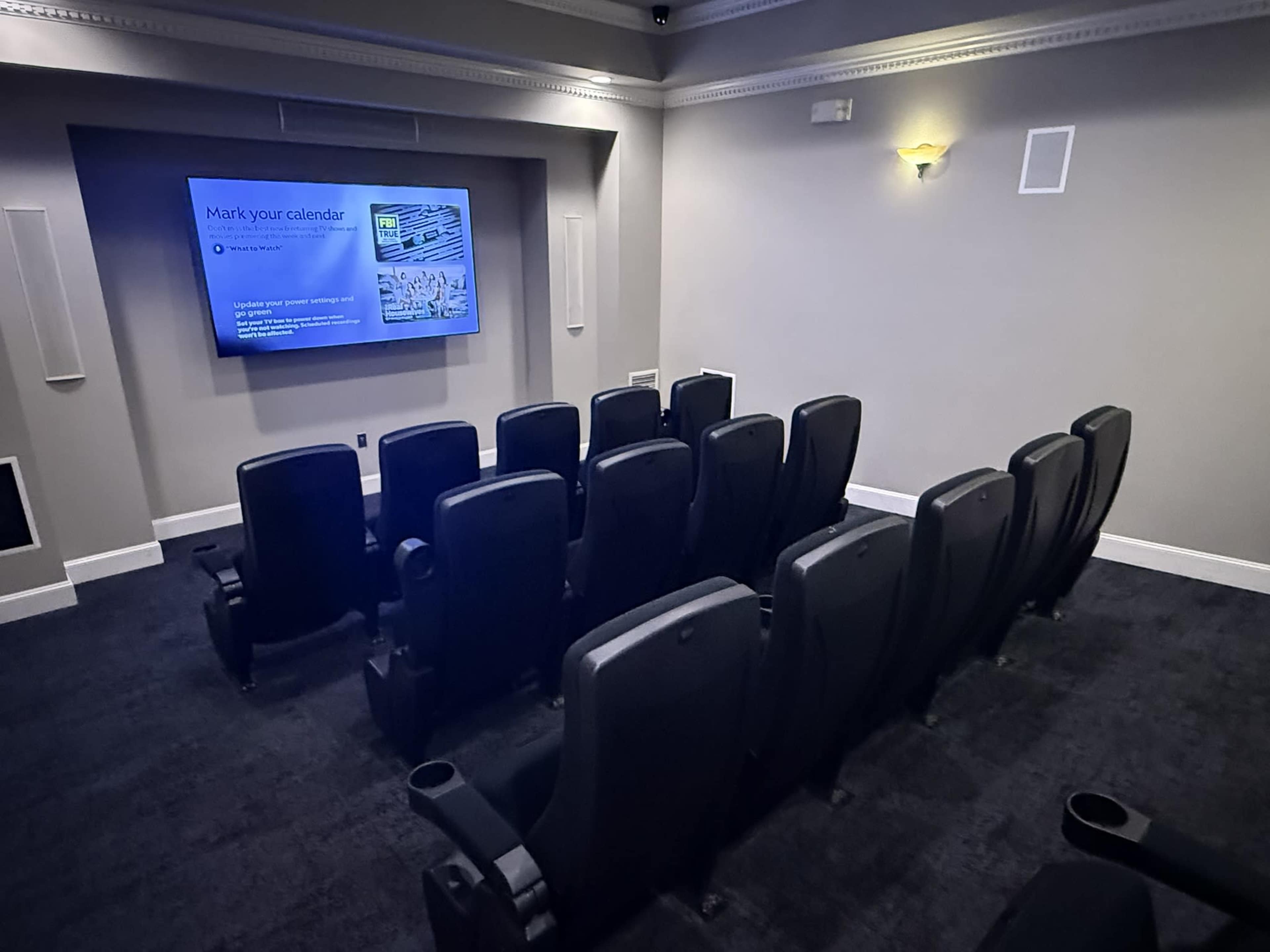 The image shows a small theater room with black seating arranged in rows facing a screen on the wall.