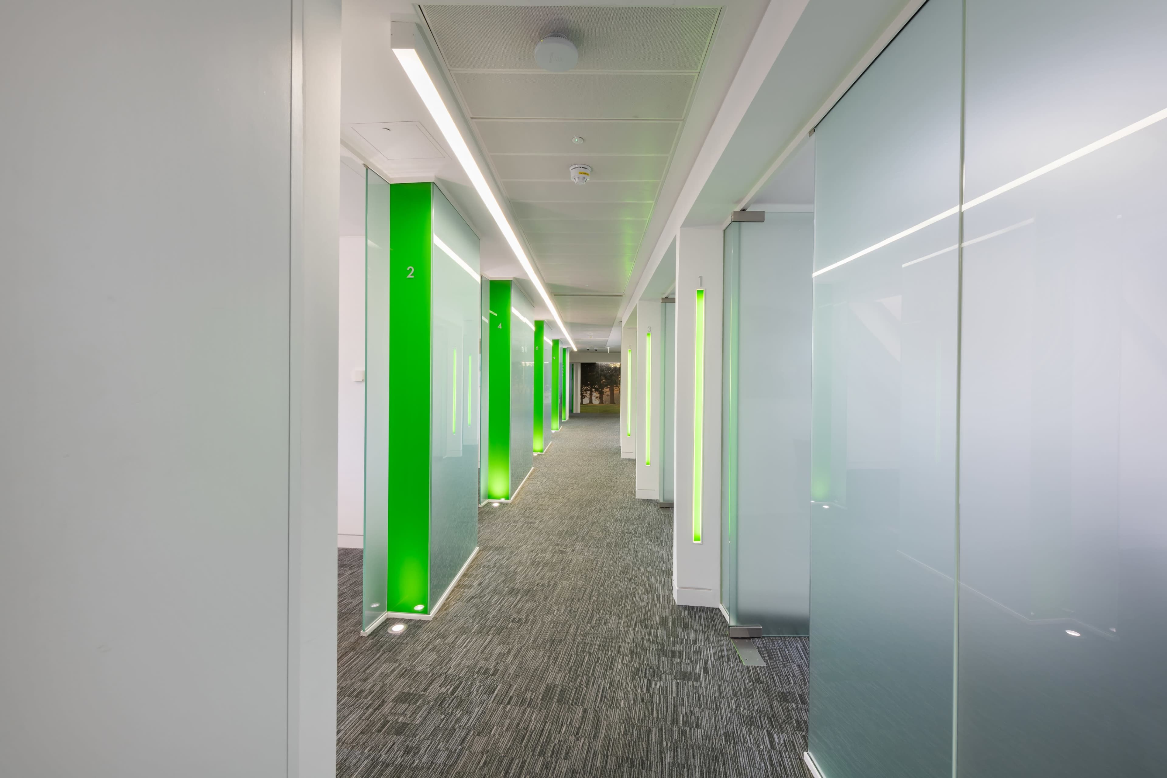 The image shows a modern office corridor with glass-walled rooms, bright green accents, and a carpeted floor.