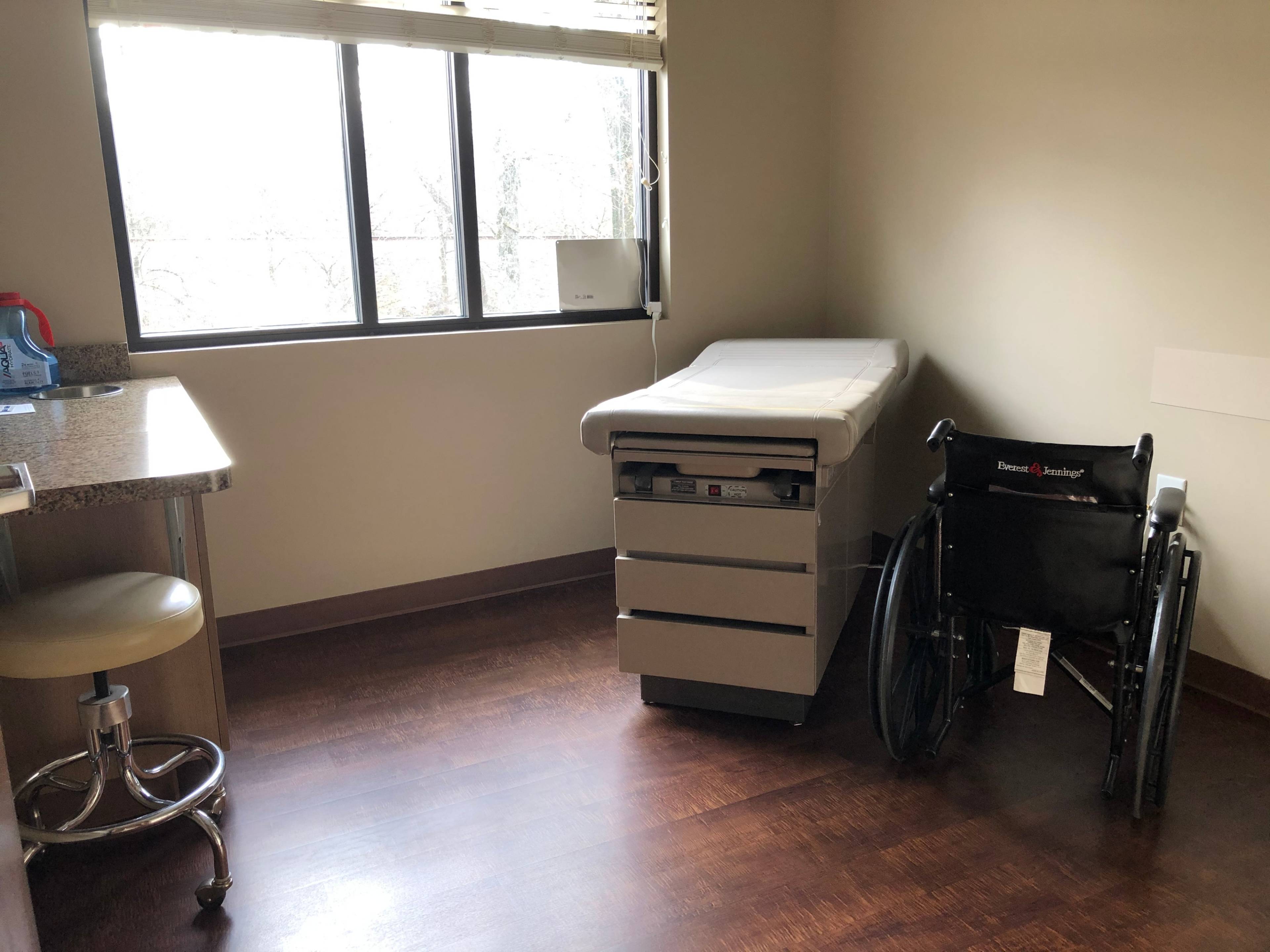 A medical examination room featuring an examination table, a wheelchair, and a window with natural light.