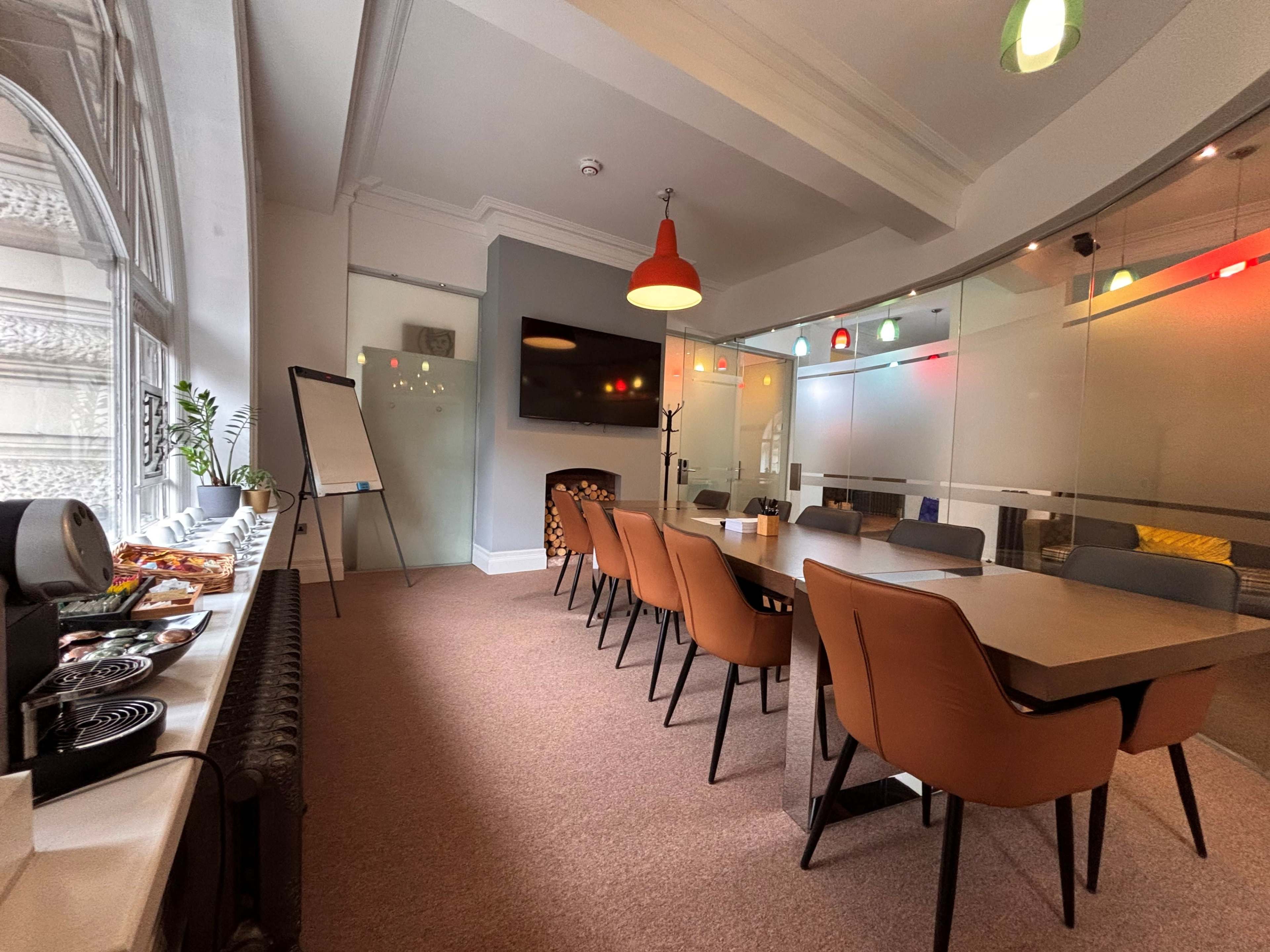 The image shows a modern conference room with a long table surrounded by brown chairs, a large screen on the wall, and a refreshment area with snacks and a coffee maker.