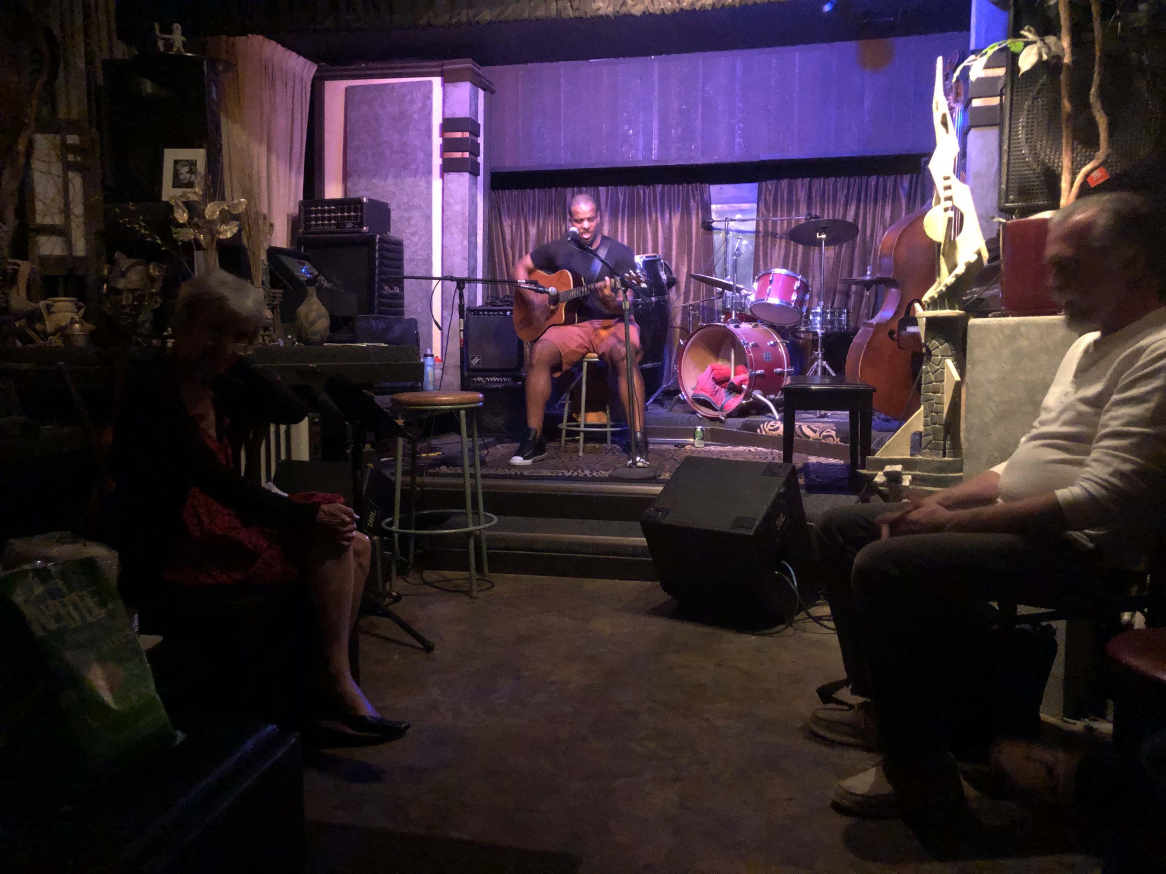 A musician plays guitar on stage while seated audience members listen in a dimly lit venue.