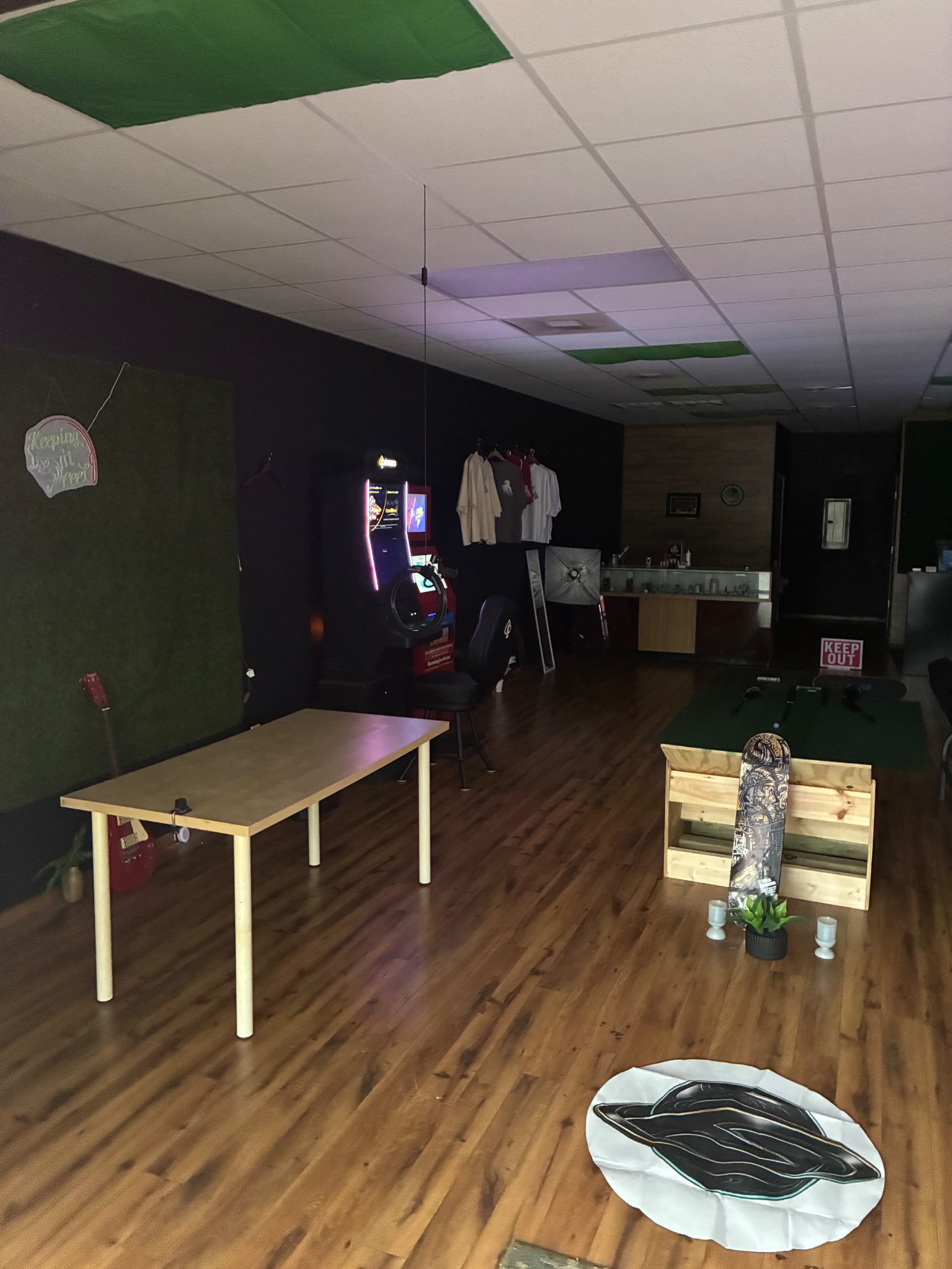 The image shows an interior space featuring a wooden table, chairs, a jukebox, and a small fenced area with a skateboard and plant decorations.