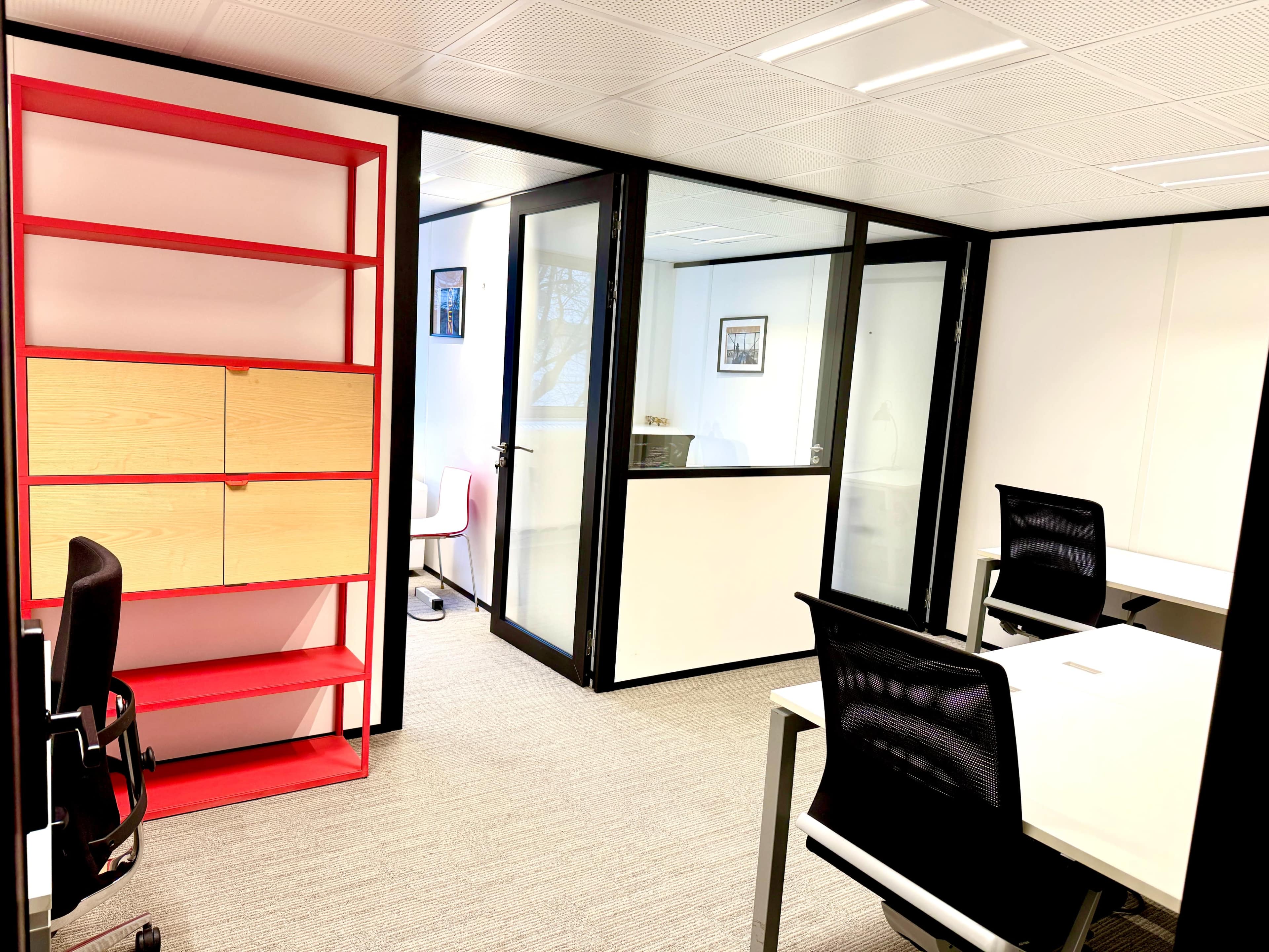 The image shows a modern office interior with a red and wooden shelving unit, two work desks, and glass-walled meeting rooms.