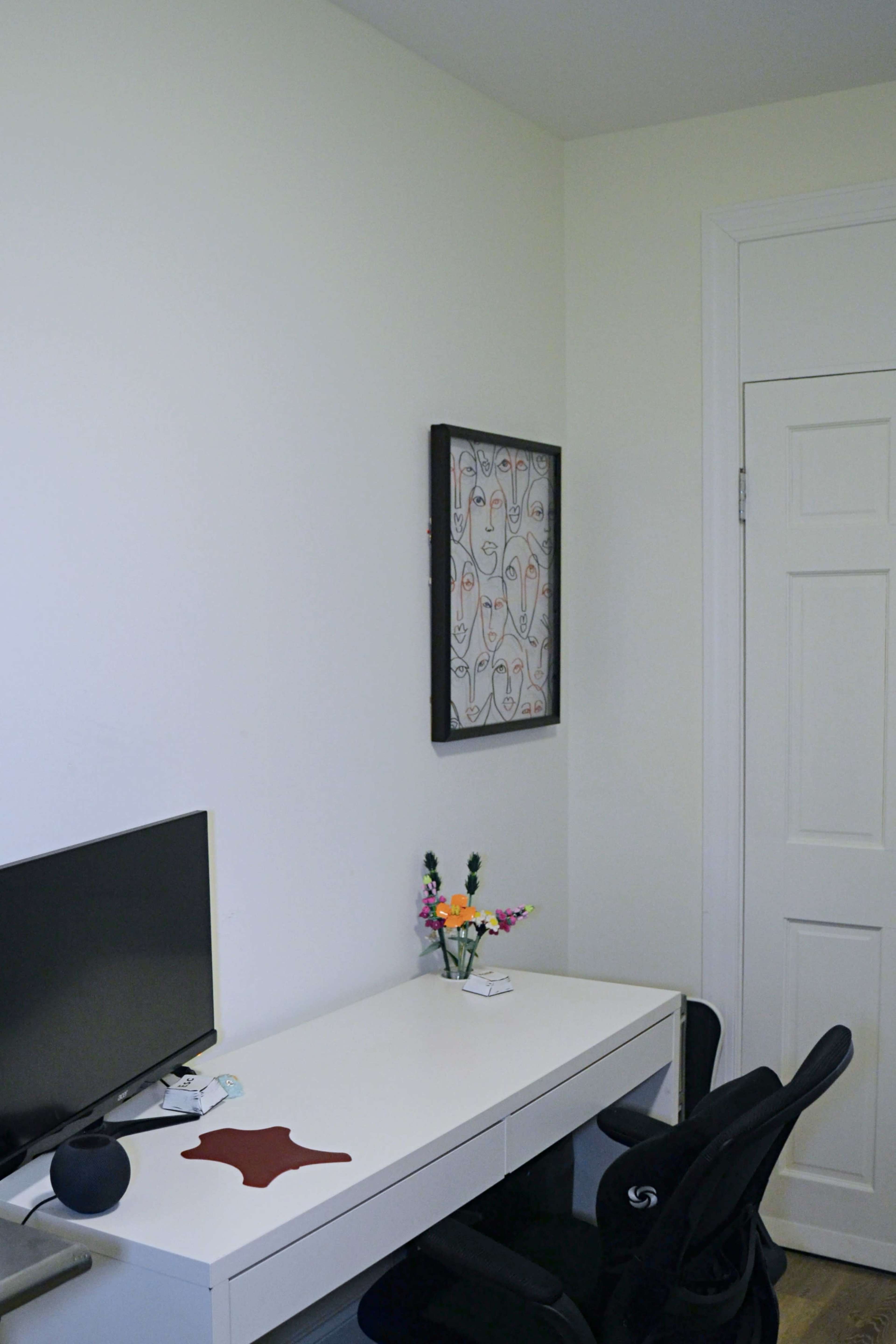 The image shows a workspace featuring a white desk with a computer monitor, a small vase of flowers, and a framed artwork on the wall beside a closed door.