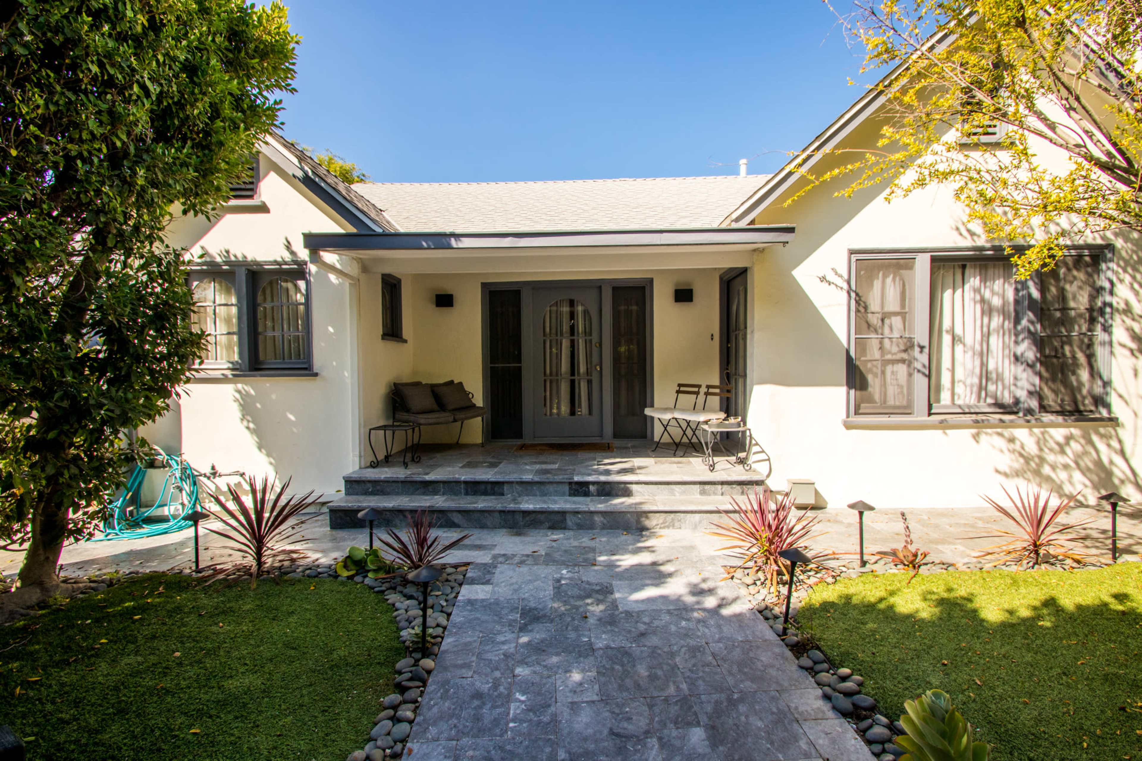 The image shows a house with a front porch, featuring stone steps leading to a door, surrounded by greenery and decorative plants.