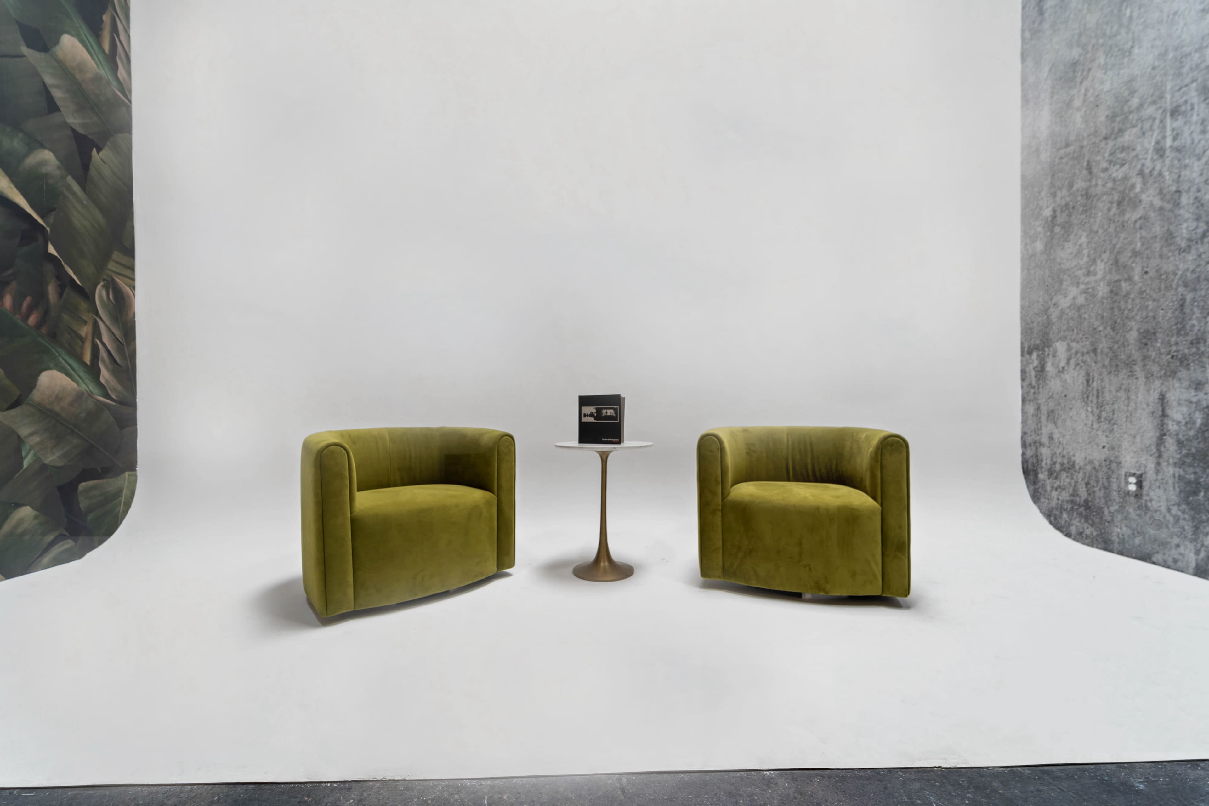 Two green velvet chairs are positioned facing each other across a small round table in a minimalist studio with a plain white background.