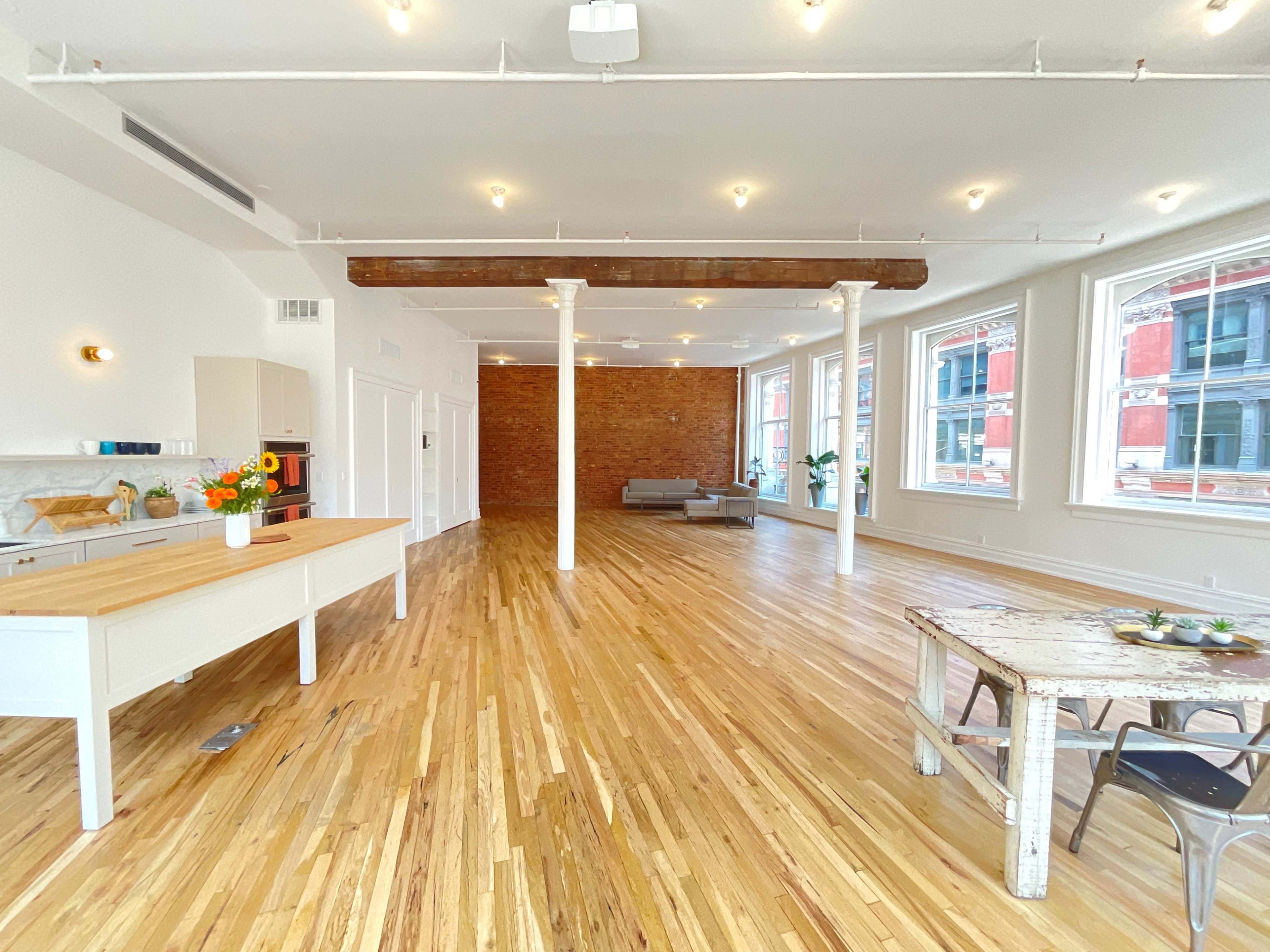 The spacious interior features hardwood floors, exposed brick walls, and large windows, with a kitchen area on one side and a seating area on the other.