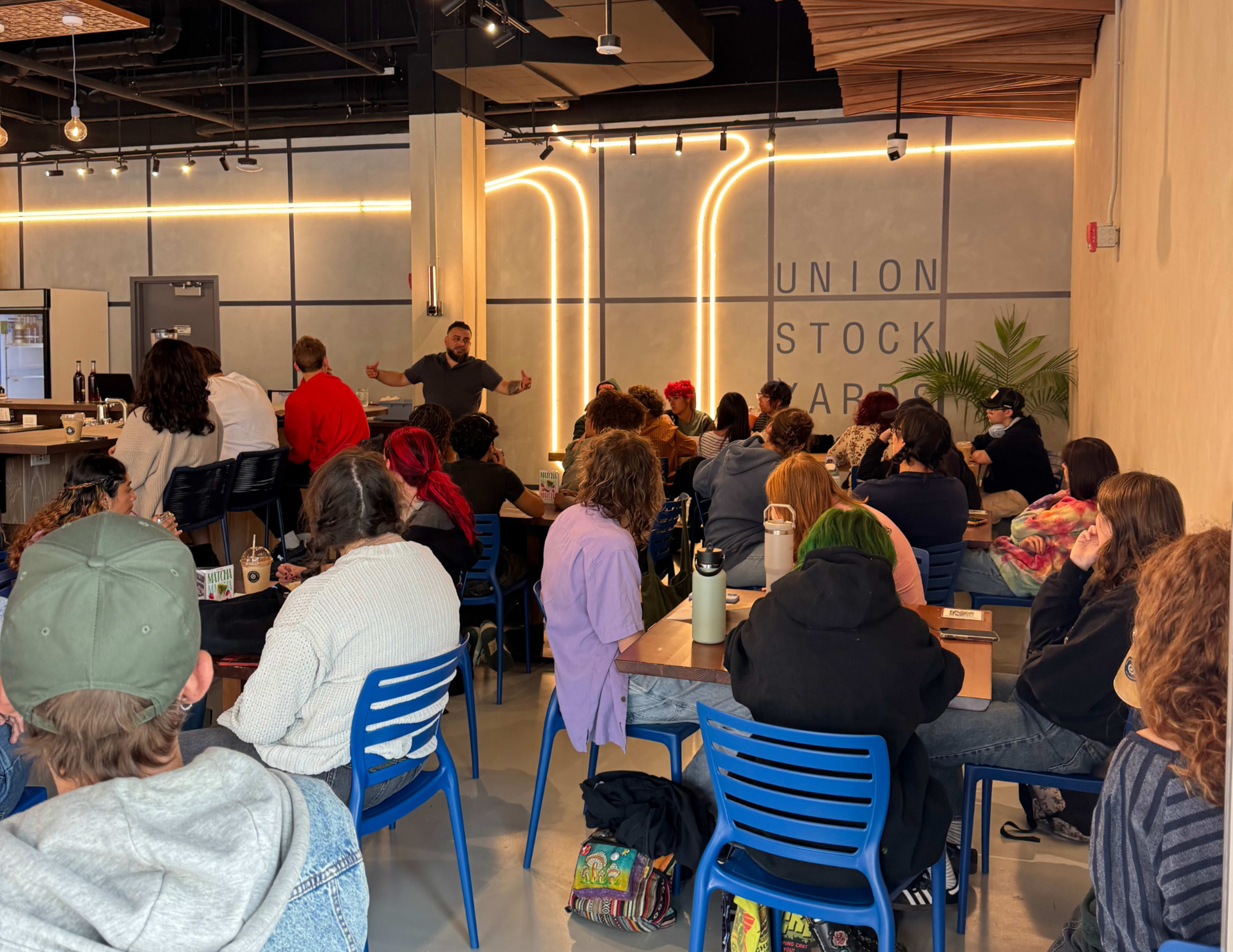 A diverse group of people attentively listens to a speaker in a modern indoor space with a café setting.