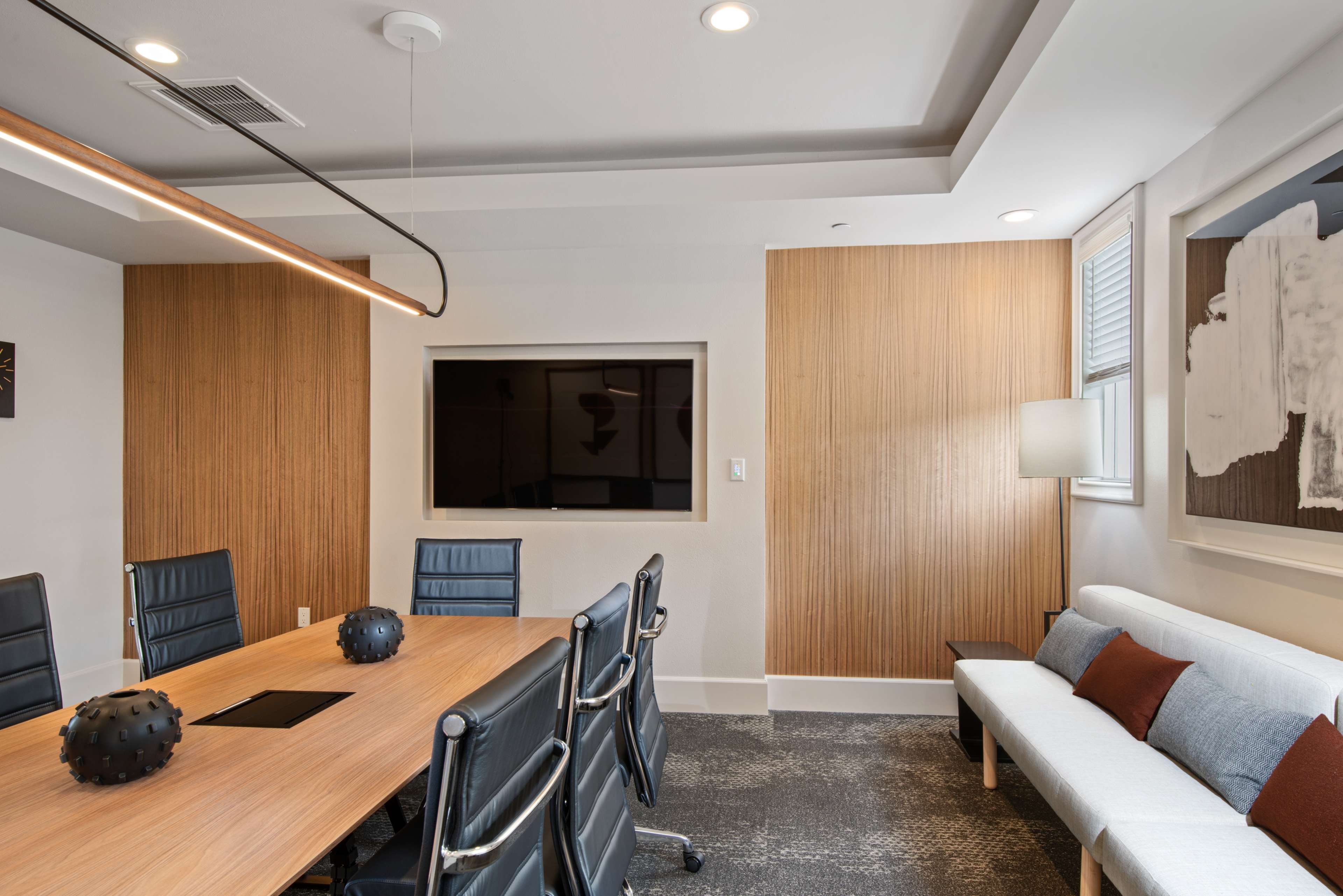 A modern conference room features a wooden table surrounded by ergonomic black chairs, with a television mounted on the wall and a minimalist couch in the corner.