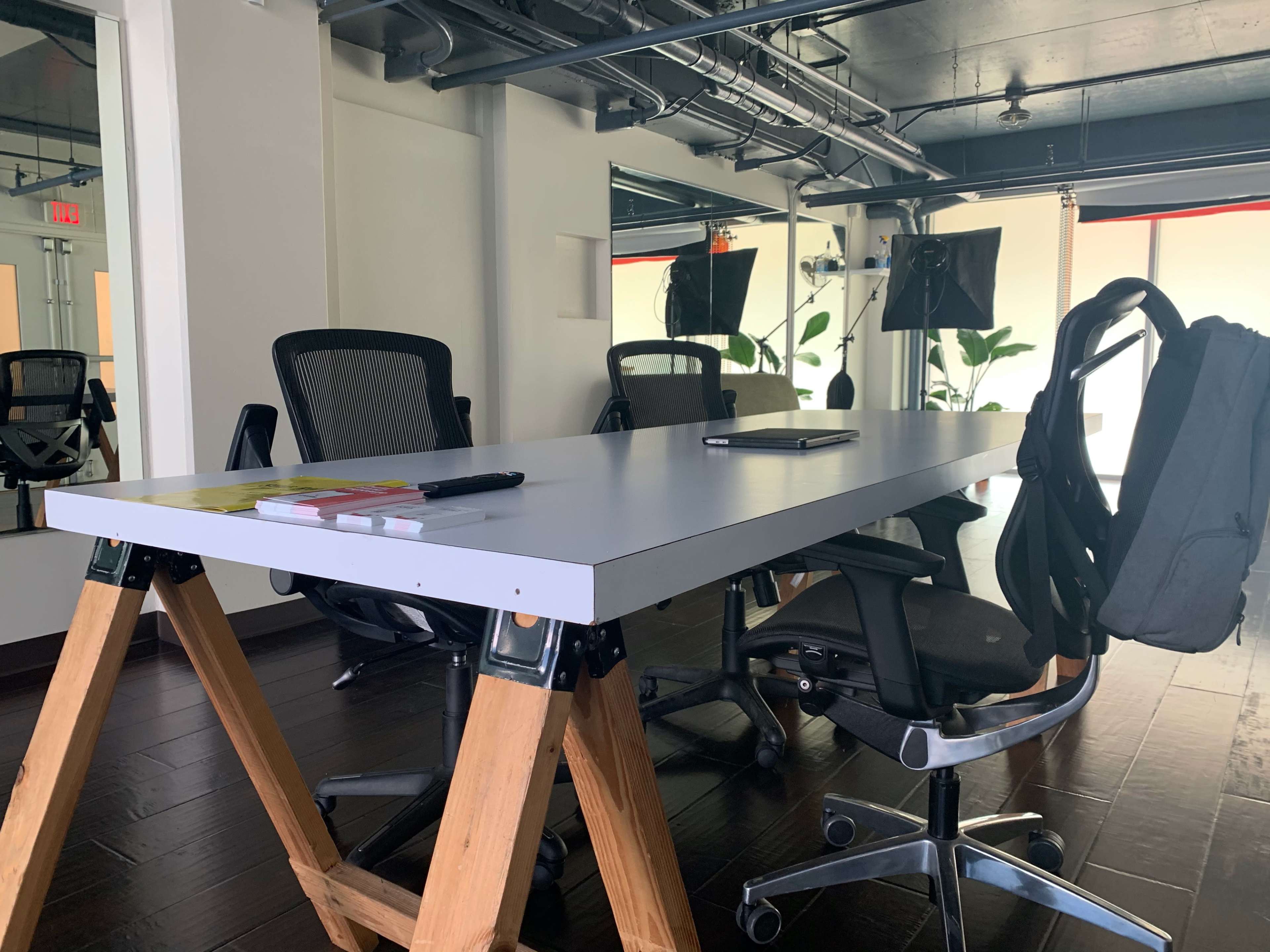 A long table with wooden legs is surrounded by several office chairs in a well-lit conference room.