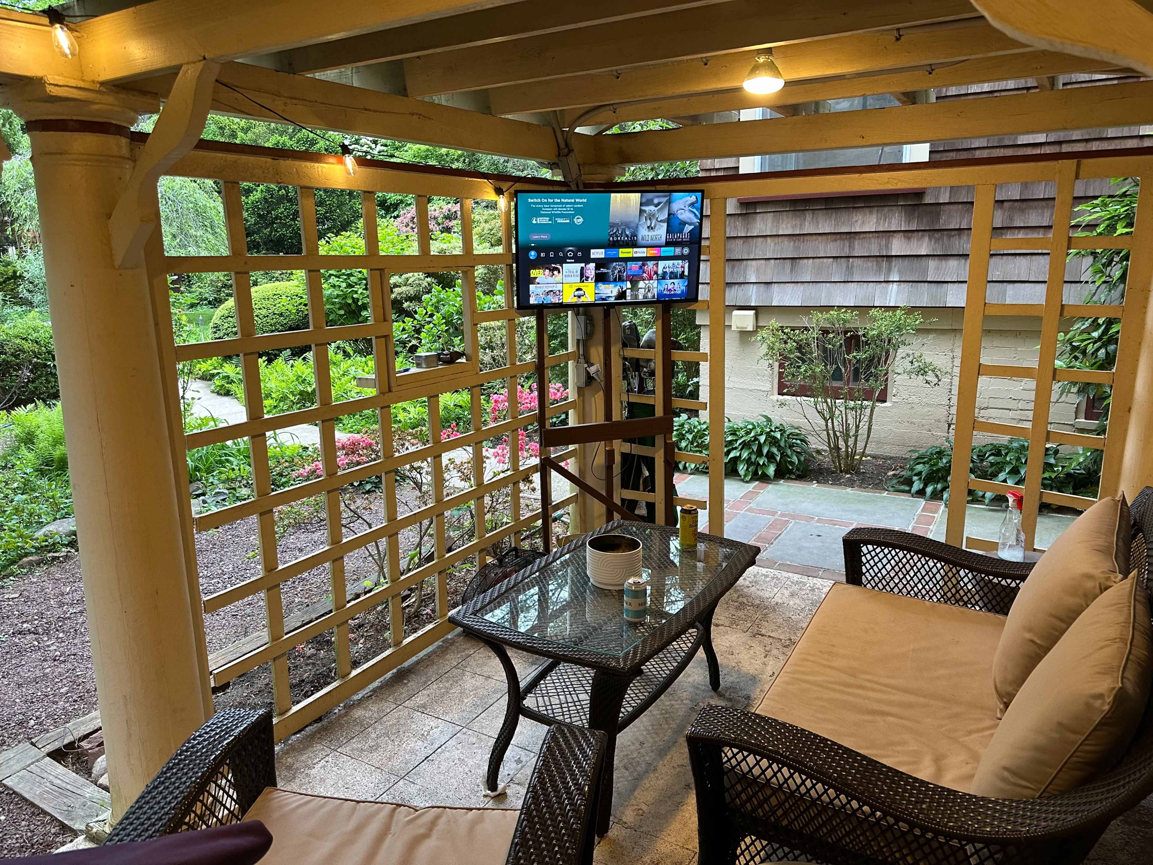 A covered patio features a seating area with two wicker chairs and a table, along with a mounted television displaying various streaming options.