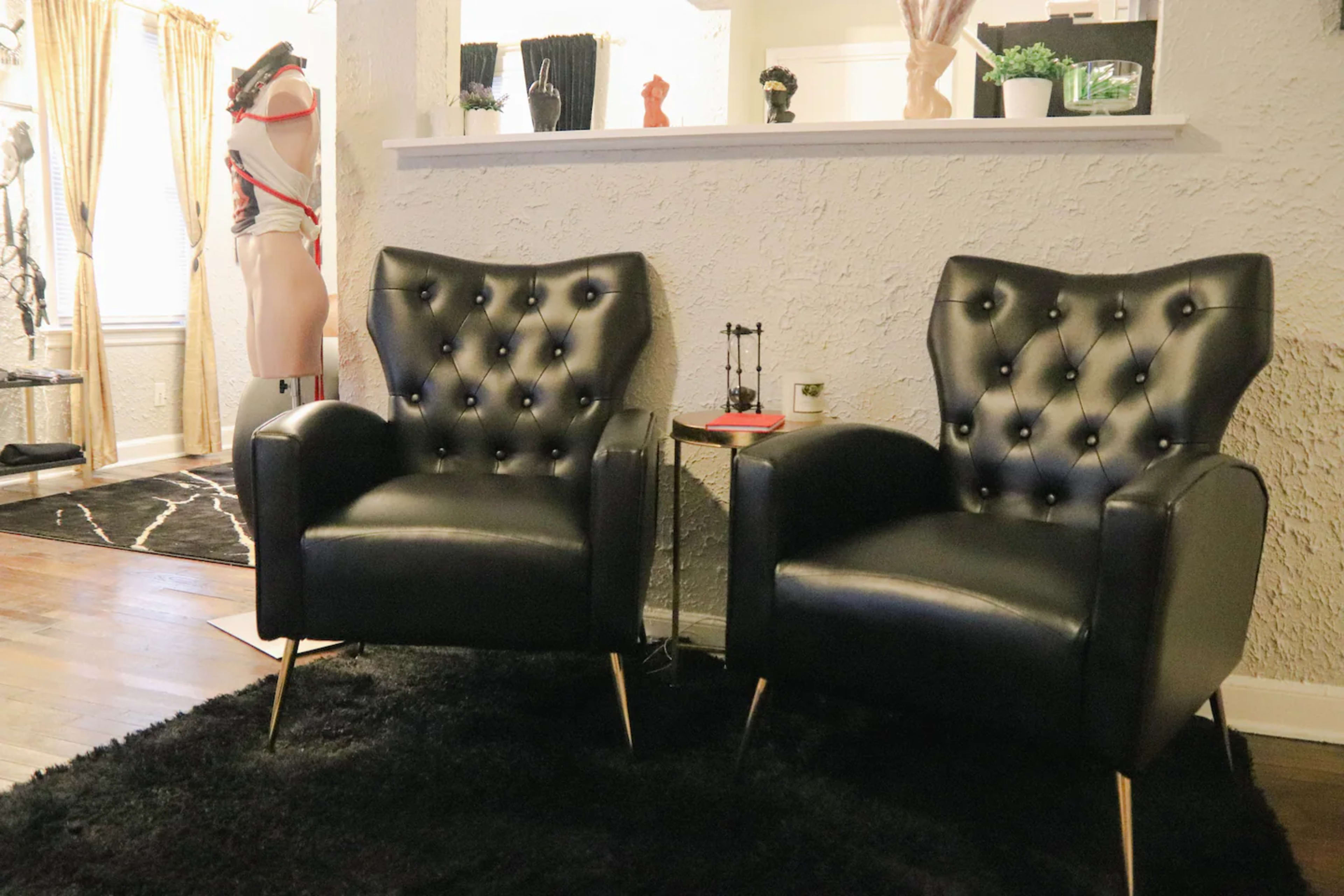 Two tufted black leather chairs with gold legs are positioned on a black rug in a room featuring various decorative items and a mannequin in the background.