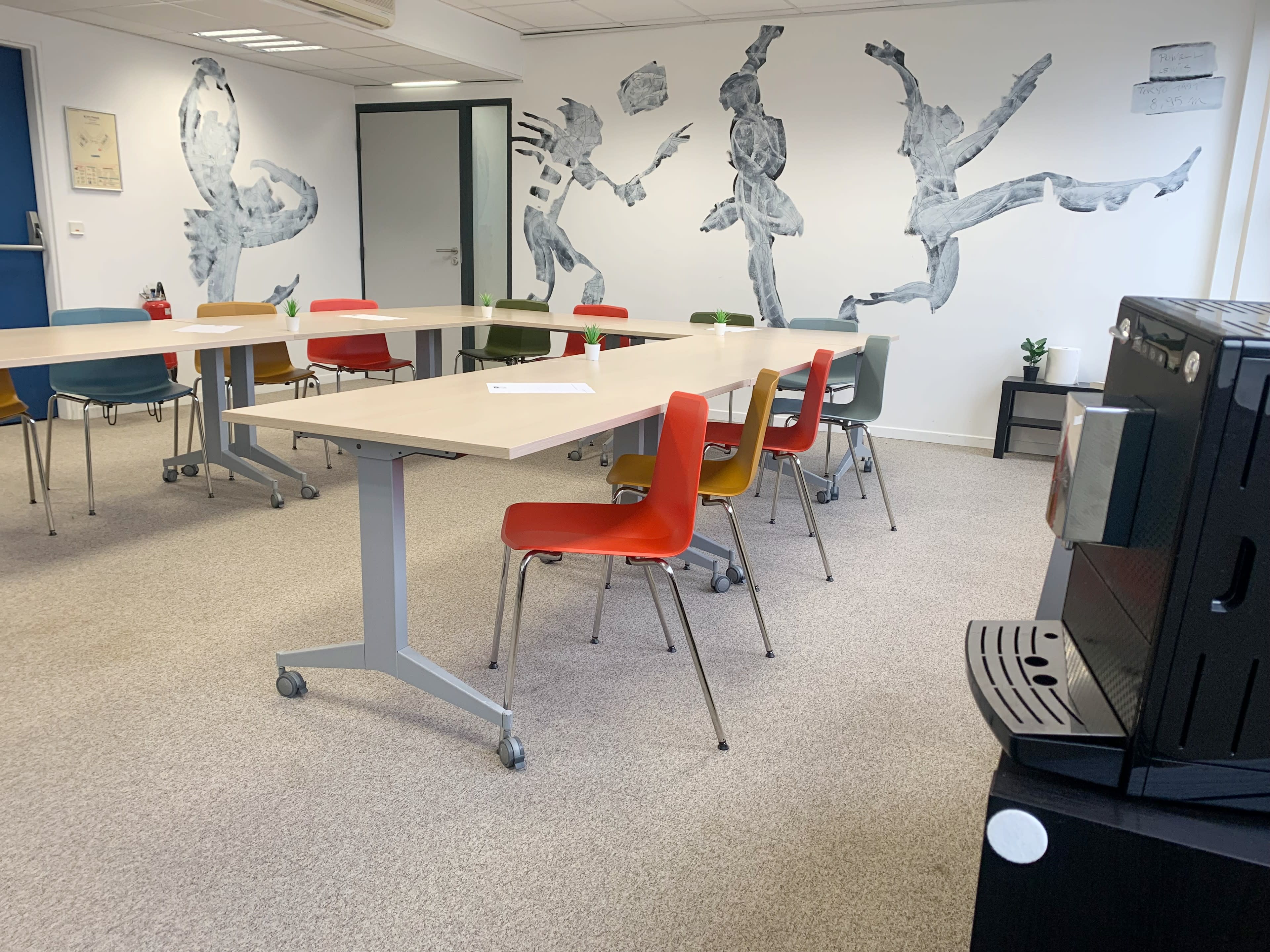 A modern workspace with several movable tables and colorful chairs, along with a large wall mural depicting figures in motion.