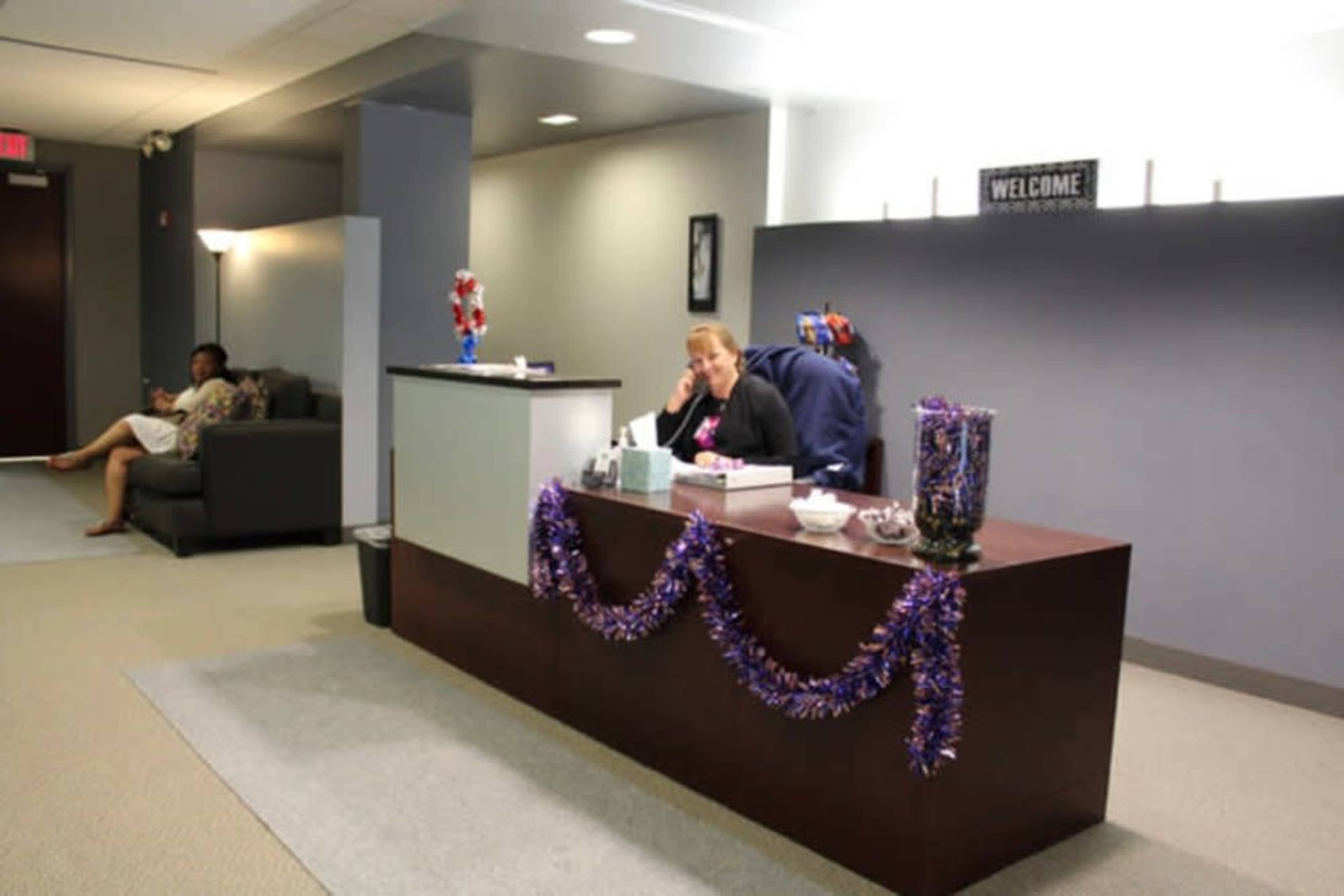 A receptionist sits at a desk adorned with purple decorations while another person relaxes on a nearby sofa in a waiting area.