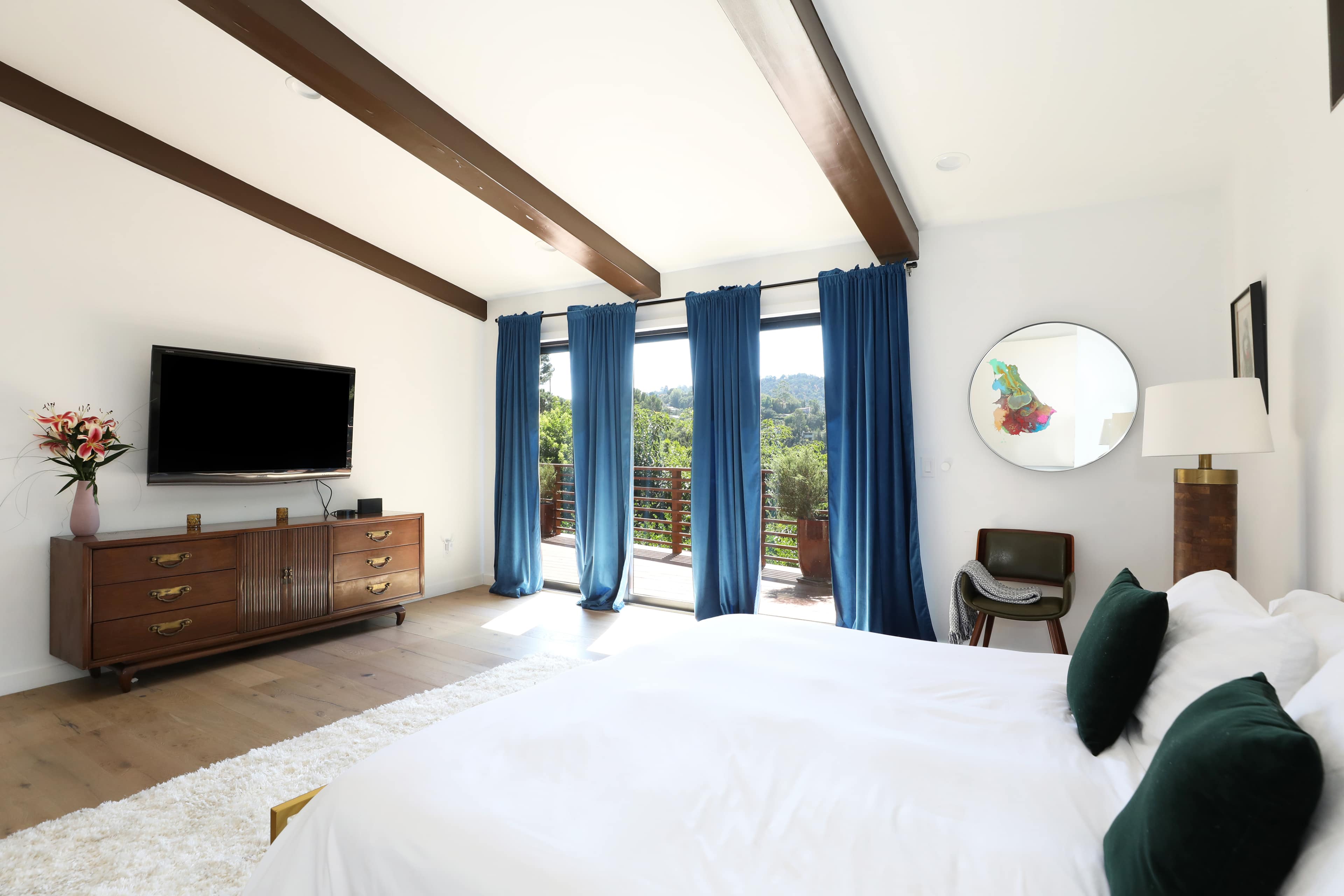 A bright bedroom features a large bed, a television mounted on the wall, blue curtains covering glass doors, and a view of a hillside through the windows.