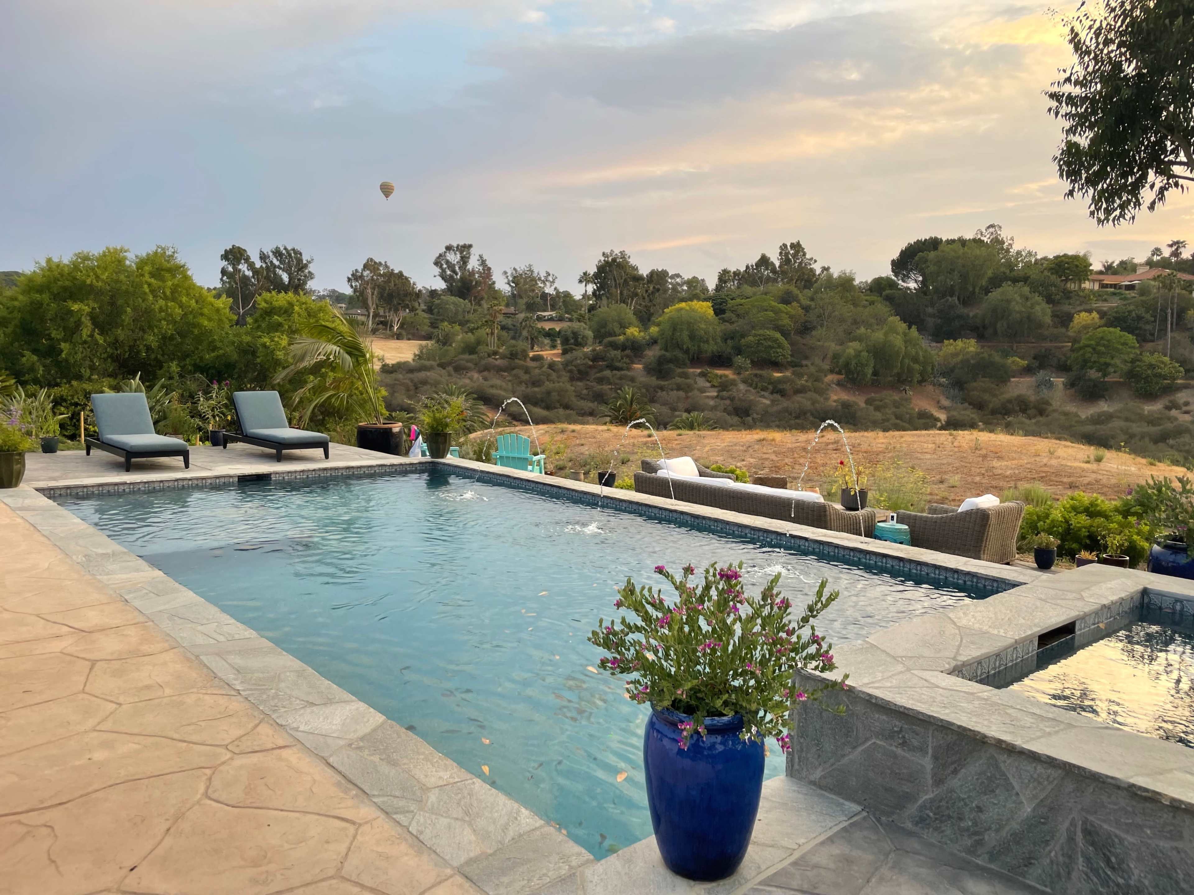 A pool area with lounge chairs, surrounded by greenery and a distant hot air balloon in the sky.