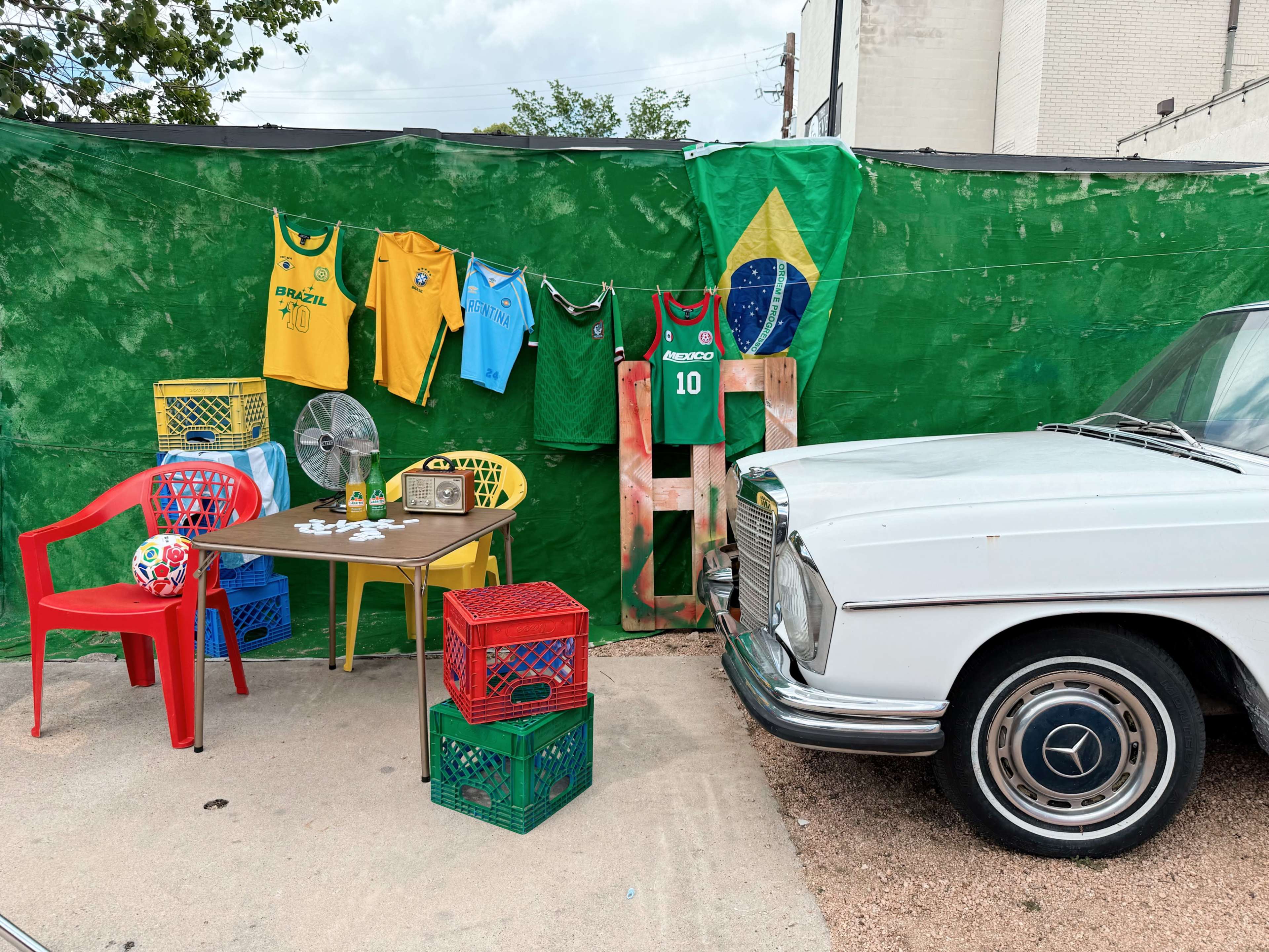 The scene features a vintage car beside colorful chairs and tables adorned with Brazilian soccer jerseys, a fan, and decorations, set against a green backdrop.