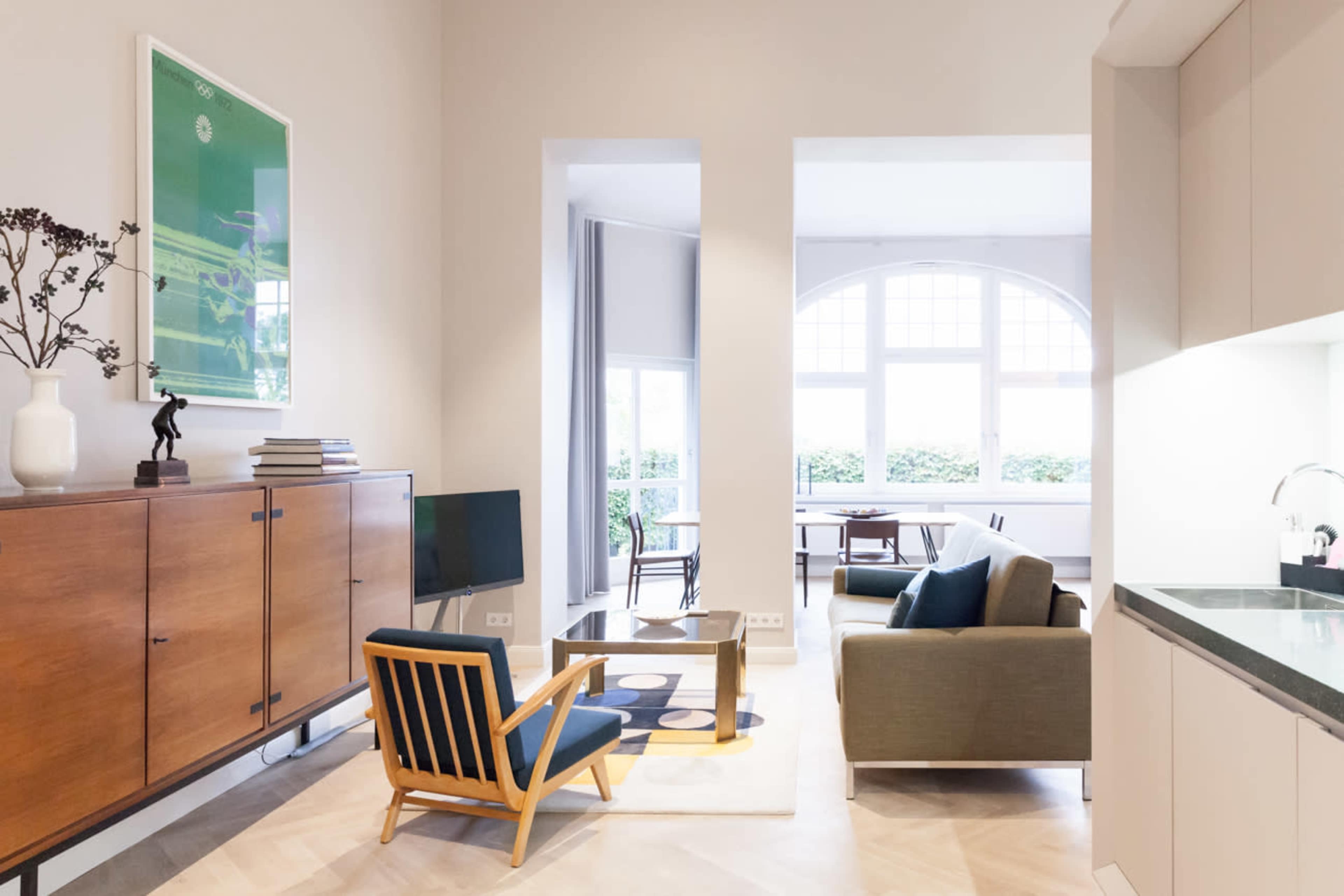 A modern living space features a wooden cabinet, a green sofa, and a mid-century chair, with large windows allowing natural light to fill the room.