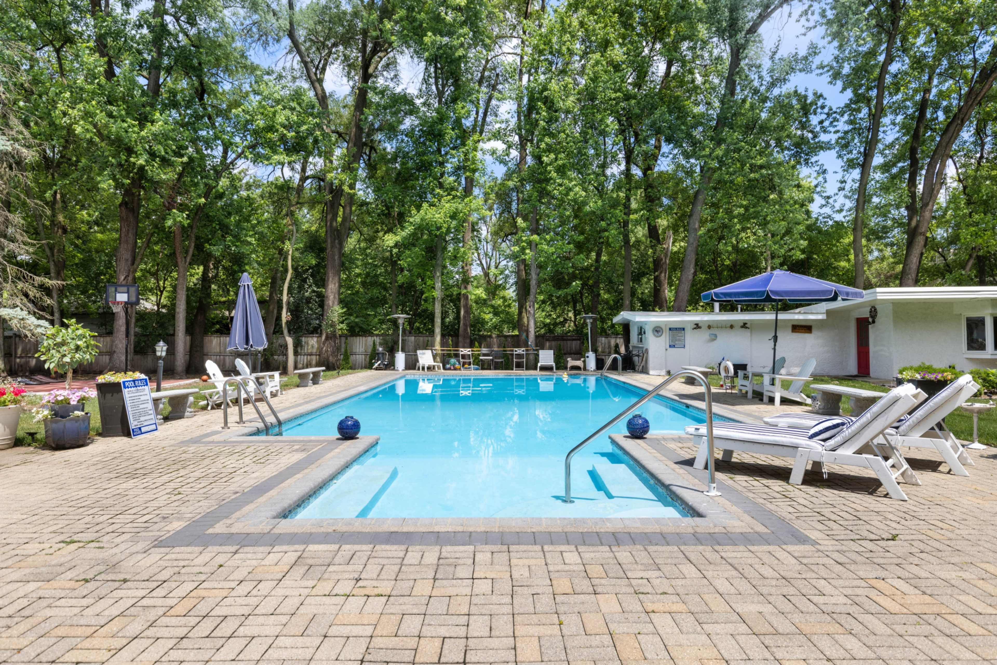 The image shows a rectangular swimming pool surrounded by lounge chairs and trees, with a blue umbrella on one side and a small building in the background.