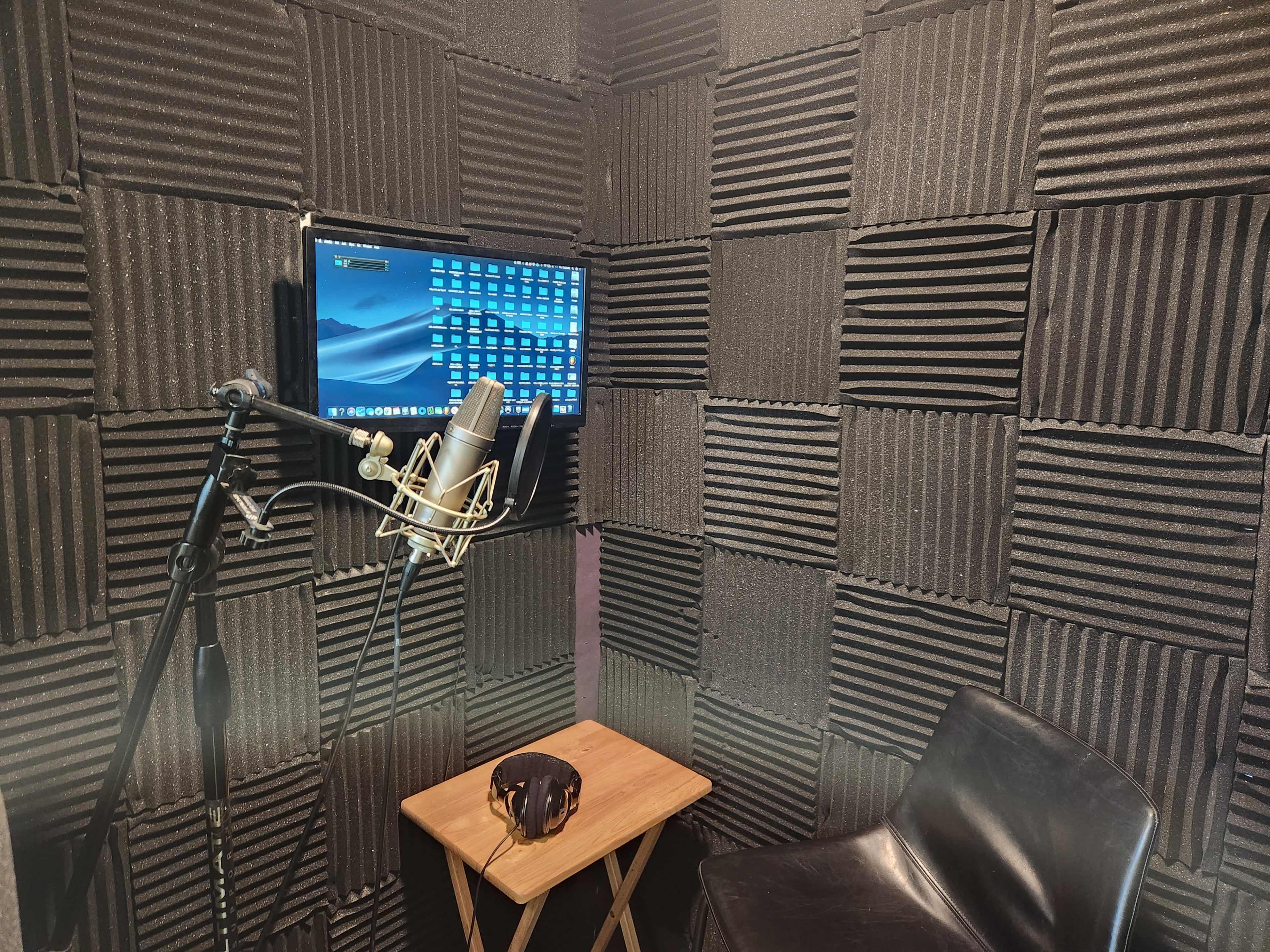 A soundproof recording studio featuring a microphone on a stand, a computer monitor mounted on the wall, a wooden table, and a black chair surrounded by acoustic foam panels.
