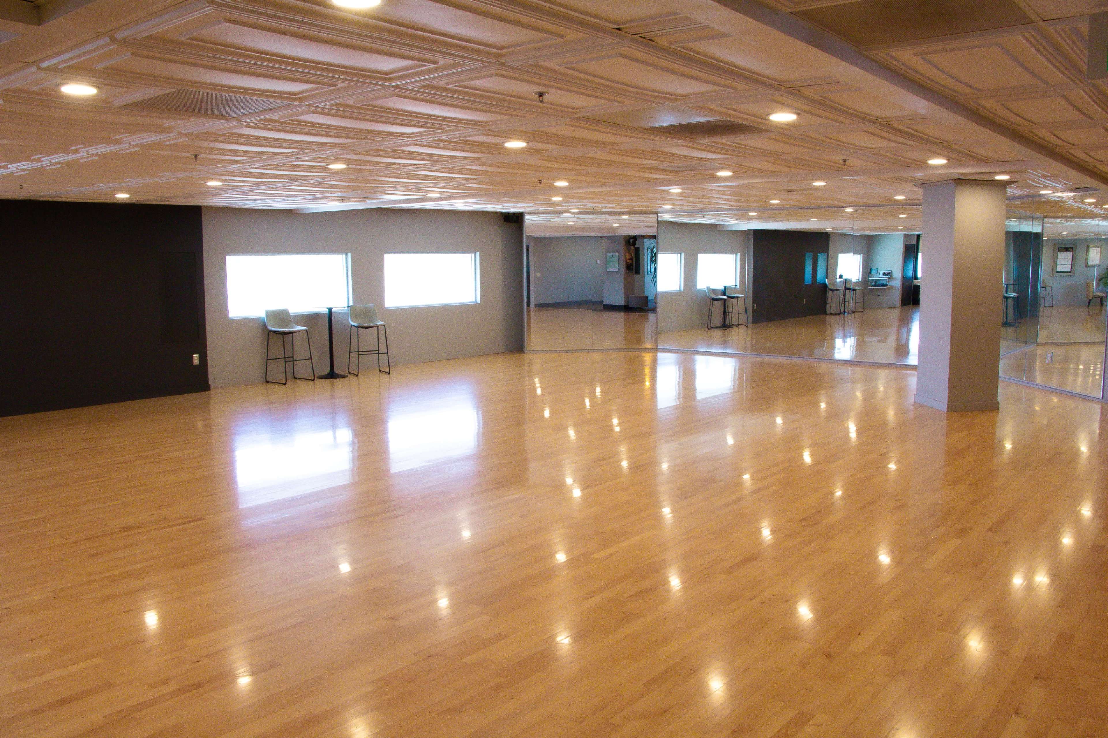The image shows a spacious, well-lit studio with hardwood flooring and large windows, featuring tall chairs against a wall and multiple mirrors reflecting the room.