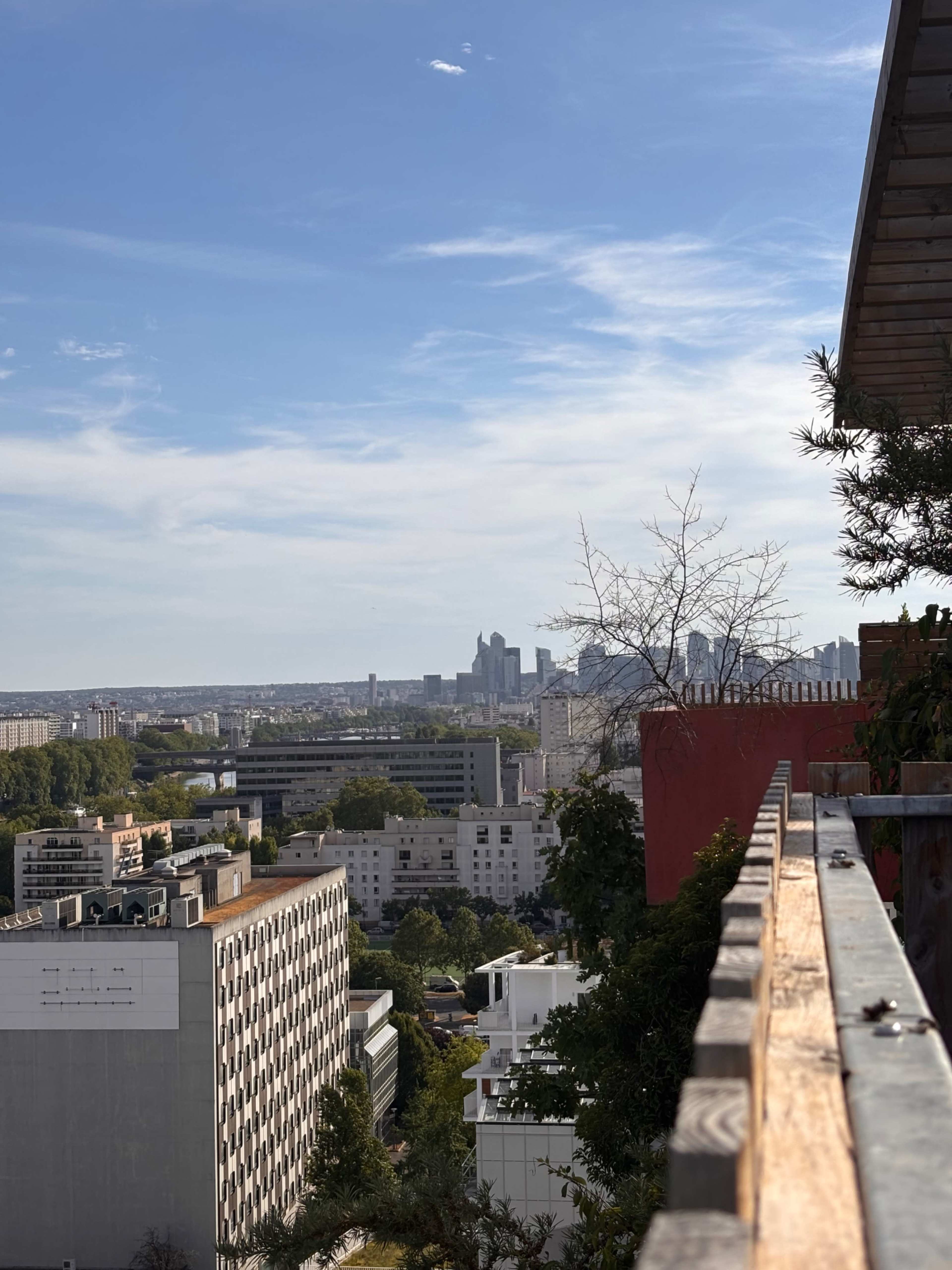 The image shows a city skyline with modern buildings under a blue sky, viewed from an elevated outdoor terrace.