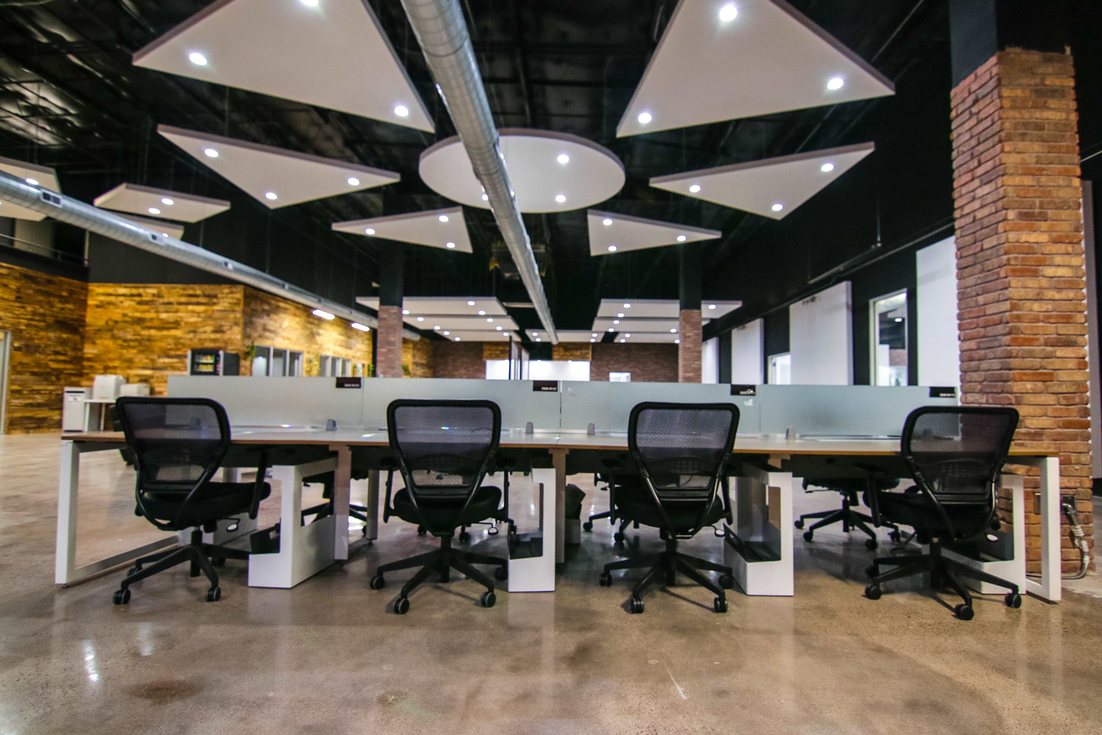 A modern office space features rows of desks with ergonomic chairs and exposed brick walls, illuminated by overhead lighting.