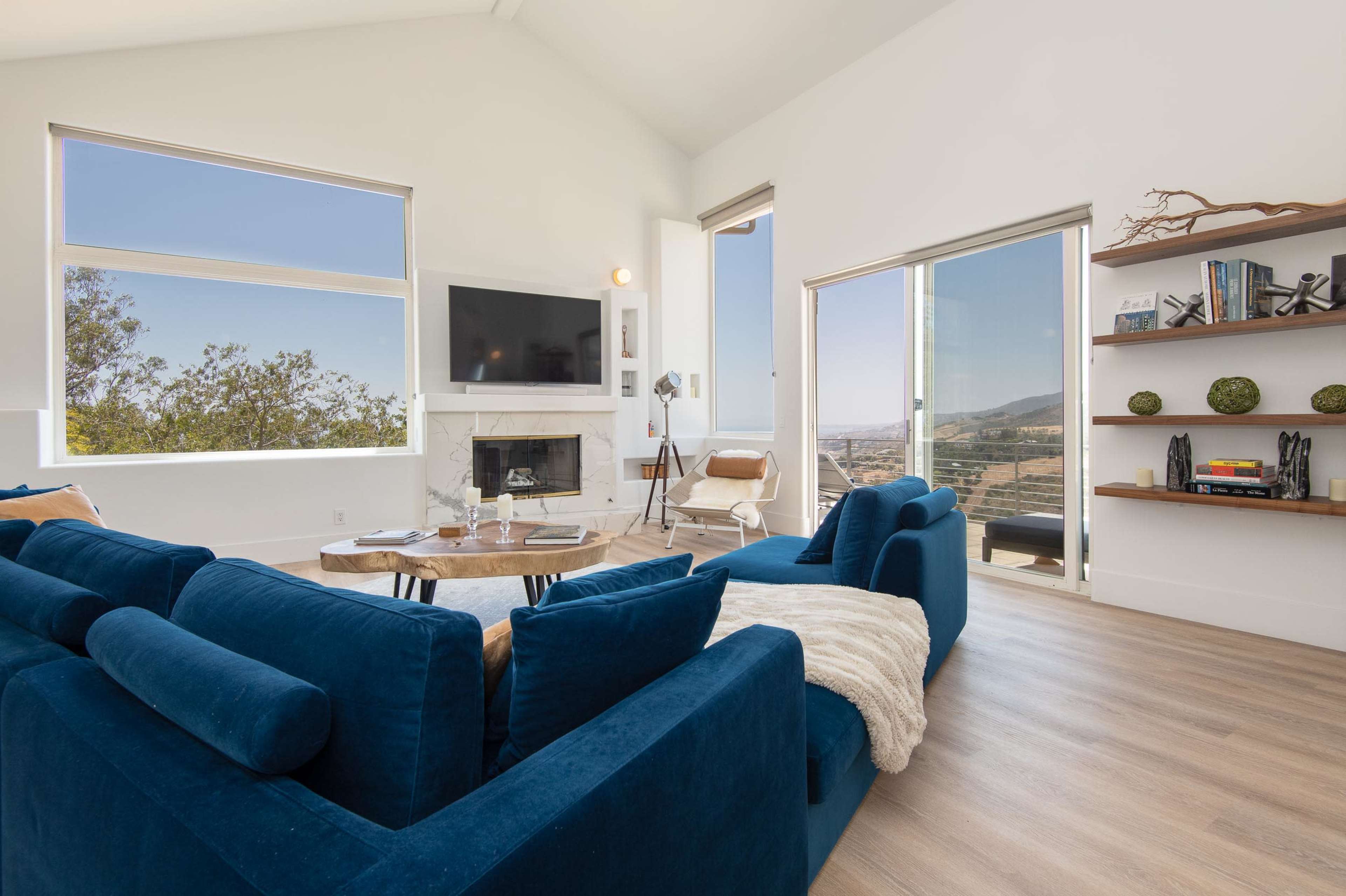 A modern living room with large windows, a fireplace, a blue sofa, and light wooden flooring.