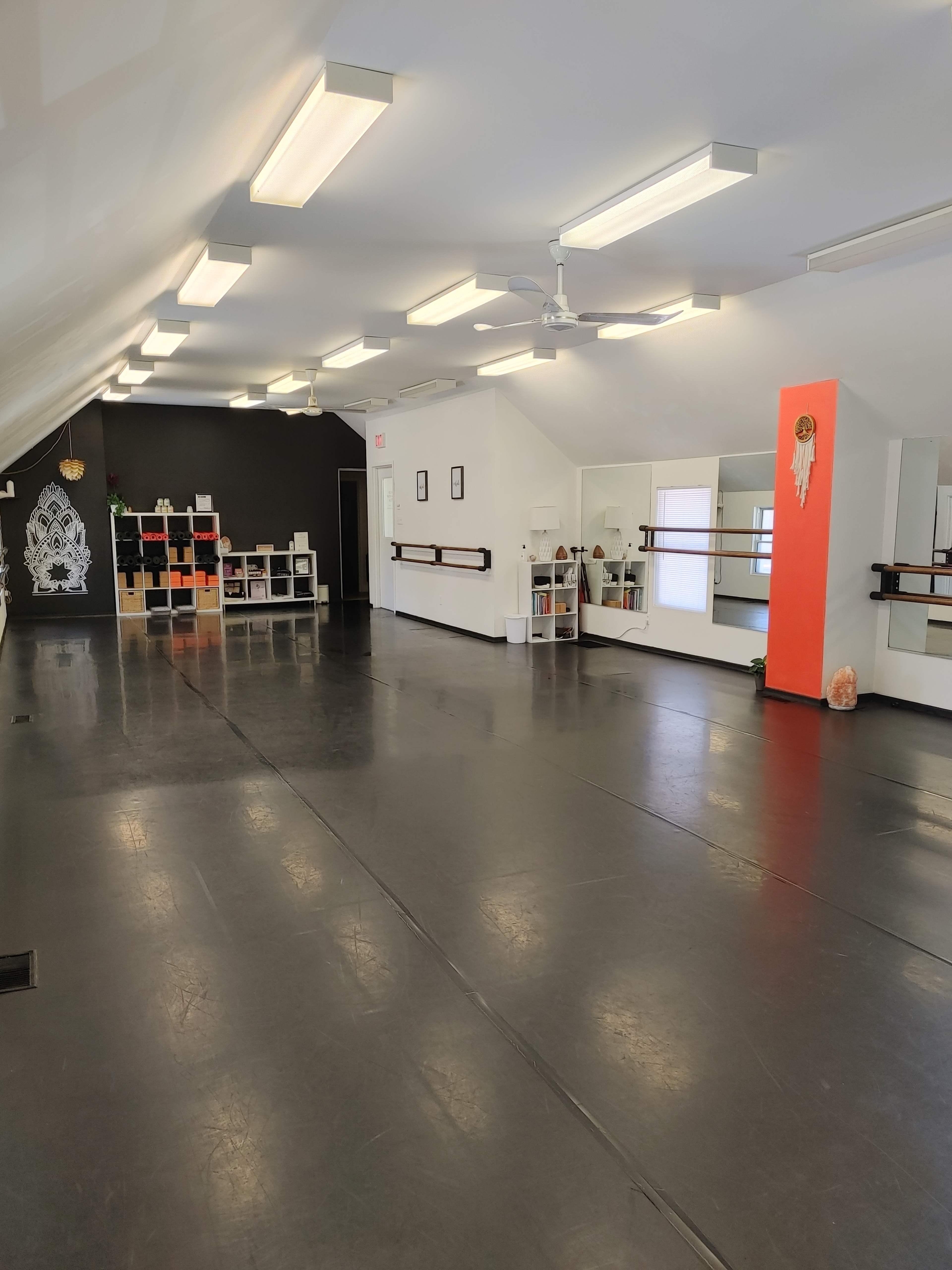 An empty dance studio with black flooring, mirrored walls, and a section featuring shelves and decorations.