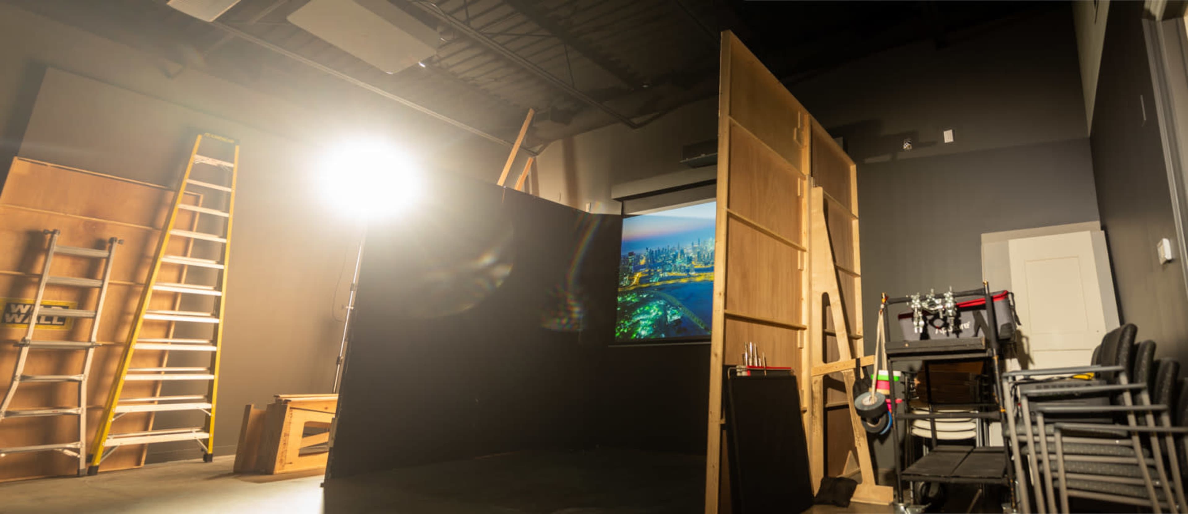 A dimly lit studio space with wooden frames and a large video projection, surrounded by a ladder and stacked chairs.